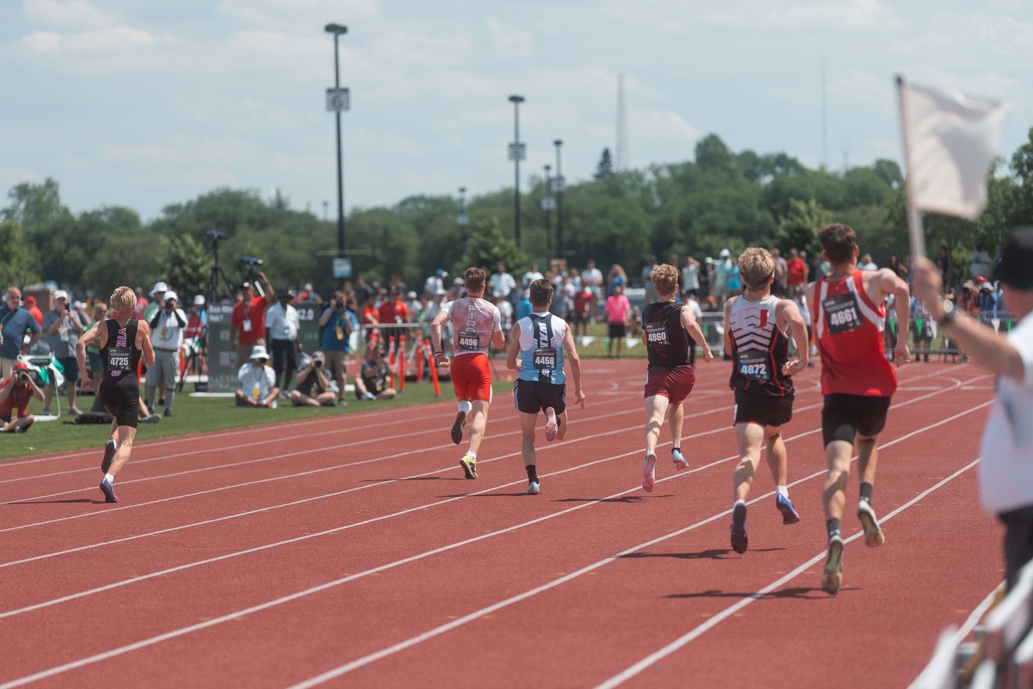 Group of young athletes running on a red outdoor track during a competitive track and field event with spectators.