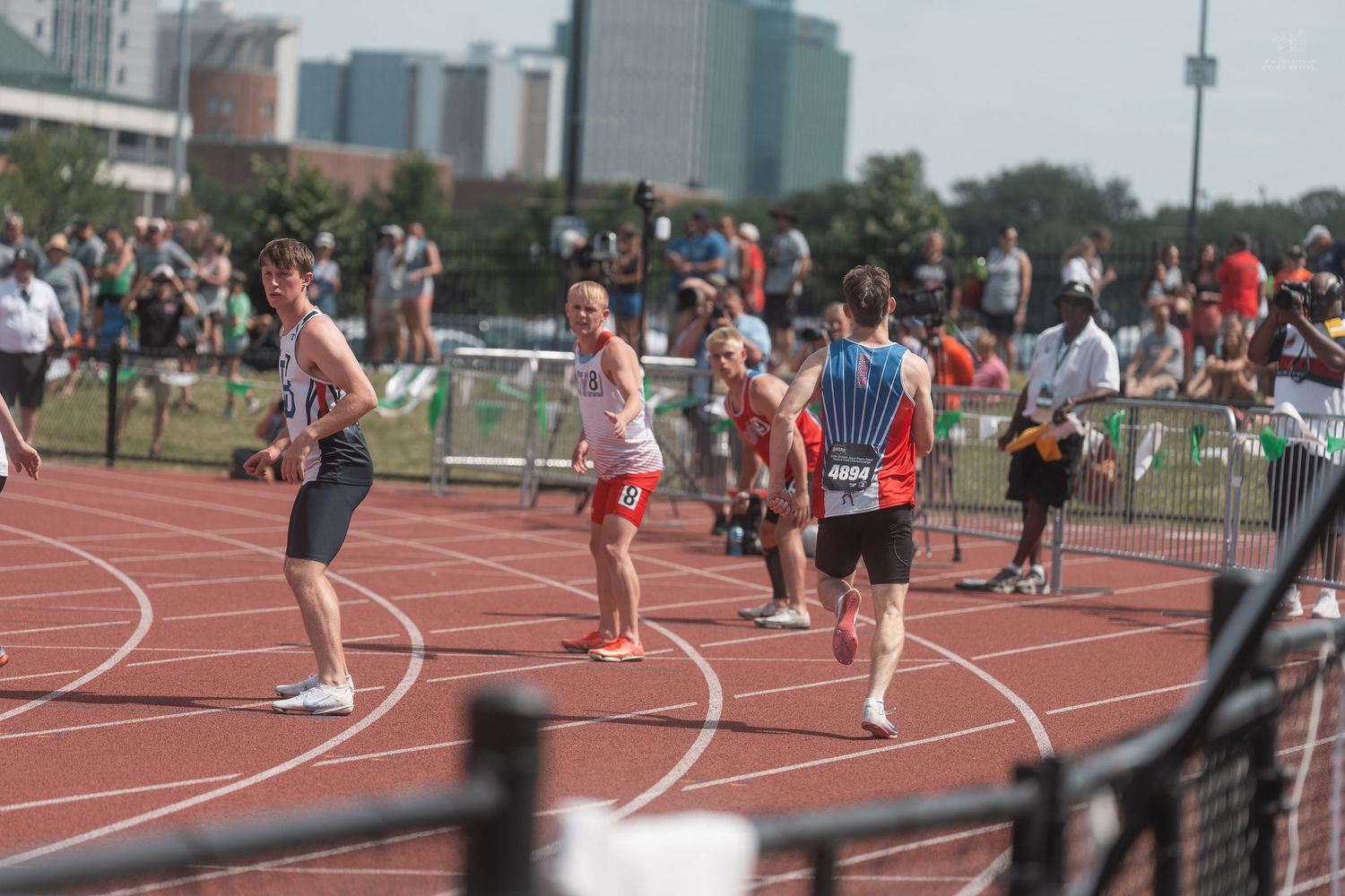Male track athletes exchange baton during outdoor relay race with crowd watching in background.