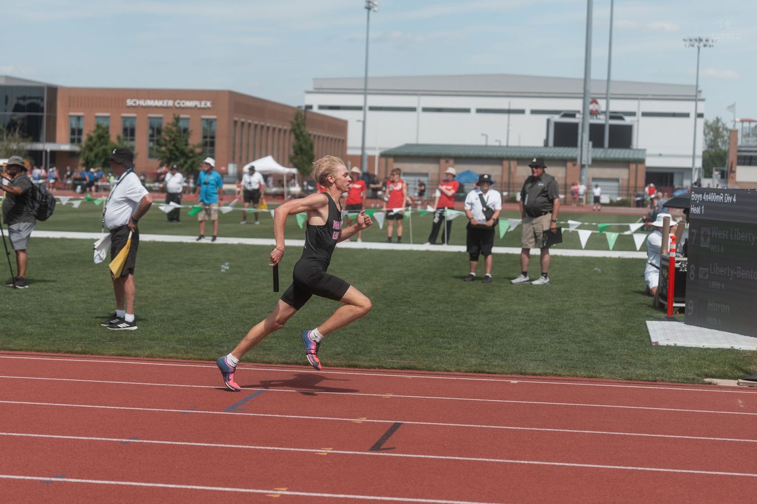 Teen athlete runs on red track at outdoor track and field competition with building and spectators in background.