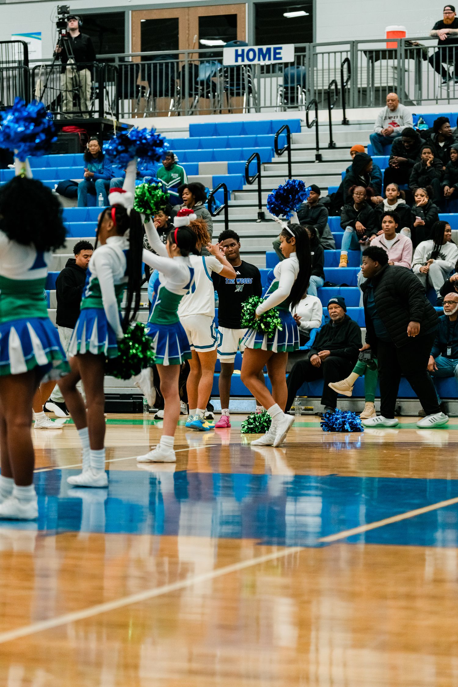 High school cheerleaders in blue and green uniforms perform routine with pom-poms in gymnasium.