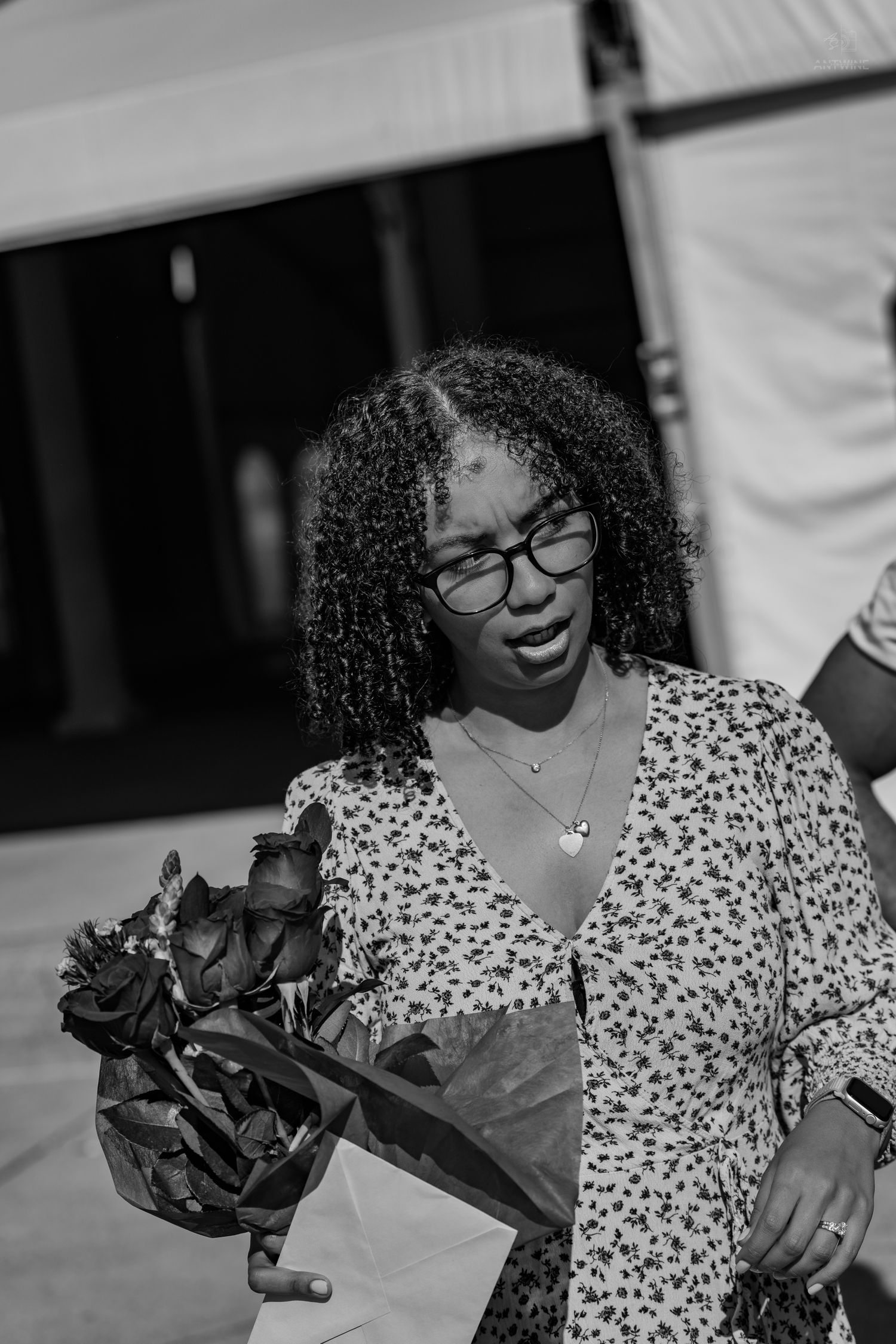 Black and white image of a young woman with glasses and curly hair holding a bouquet of roses outdoors.