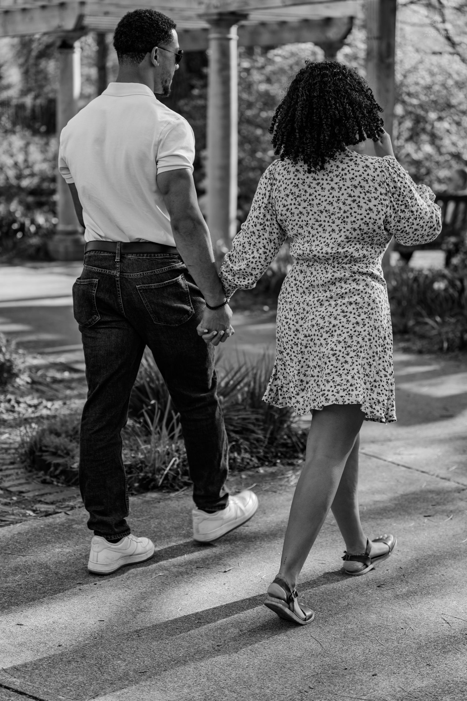 Black and white image of a couple holding hands walking along a sunlit garden pathway.