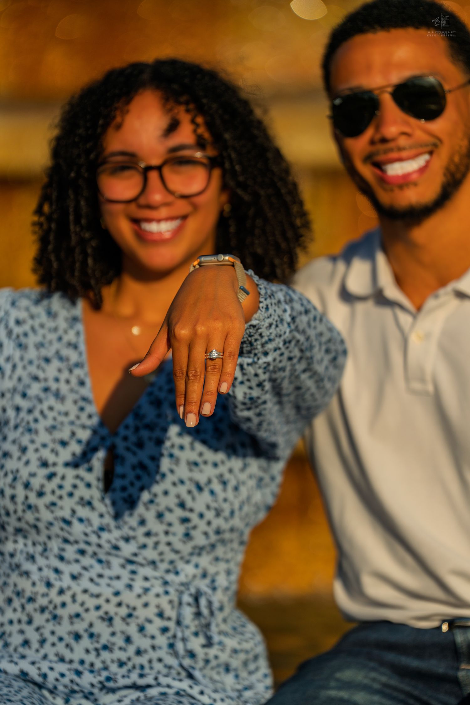 A happy couple proudly displays an engagement ring while sitting together in warm golden light.