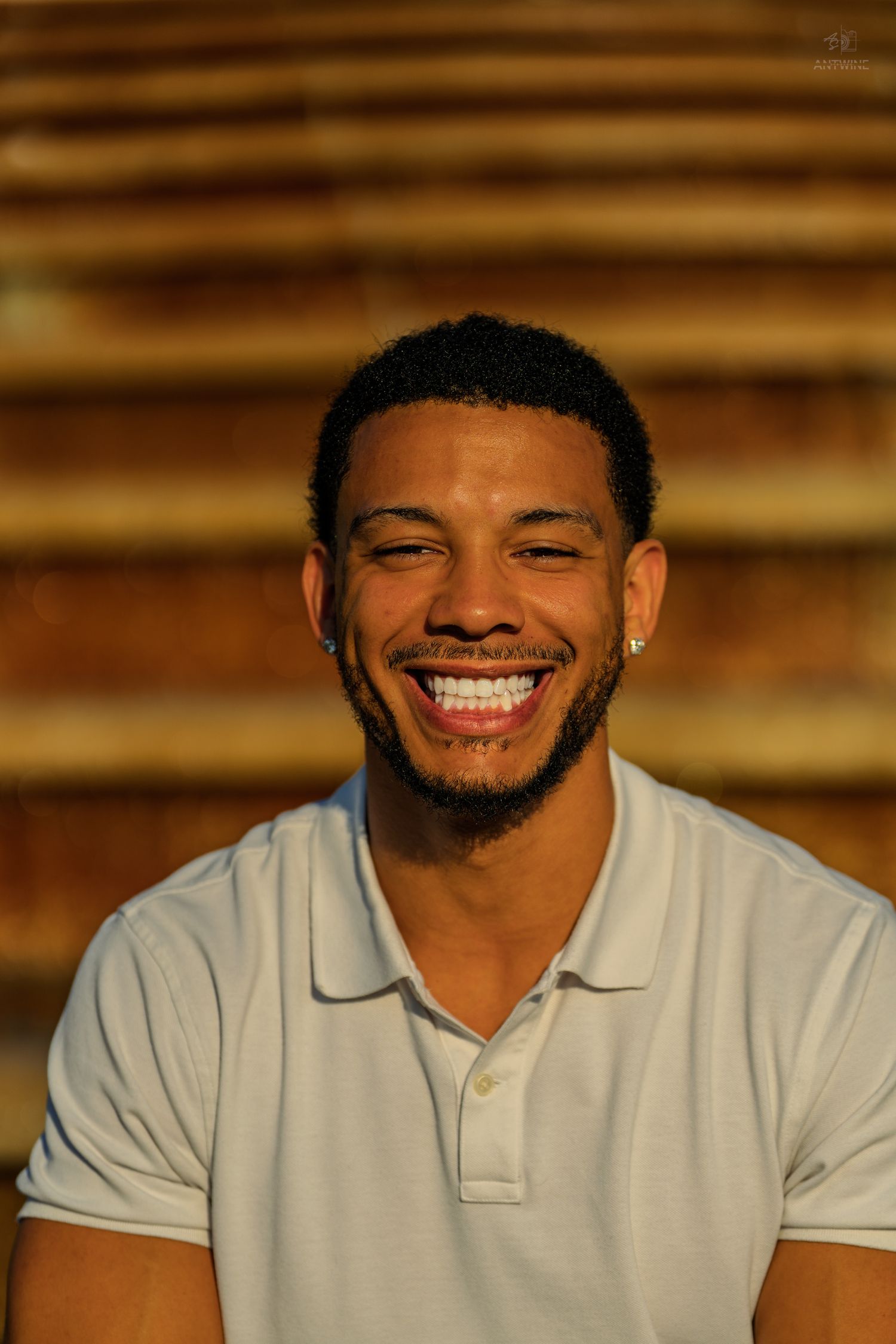 A young Black man in a white polo shirt laughs joyfully in front of warm golden stone steps.