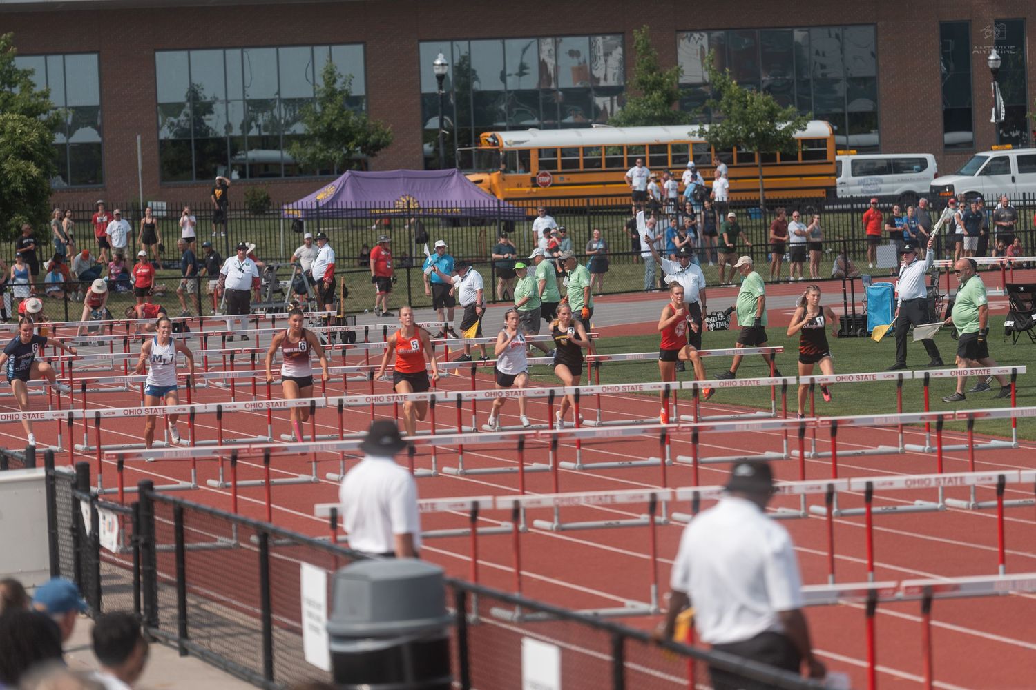 Athletes compete in hurdles race at packed outdoor track and field stadium with school bus in background.