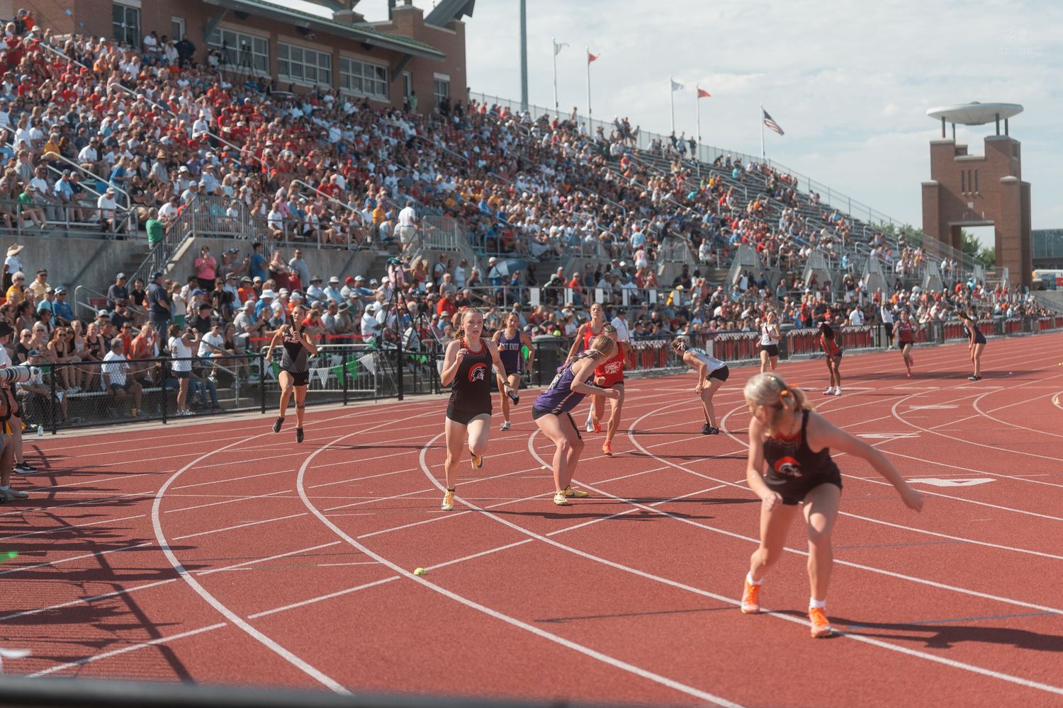 Female athletes race on a red track during a relay event at a large outdoor track and field meet.