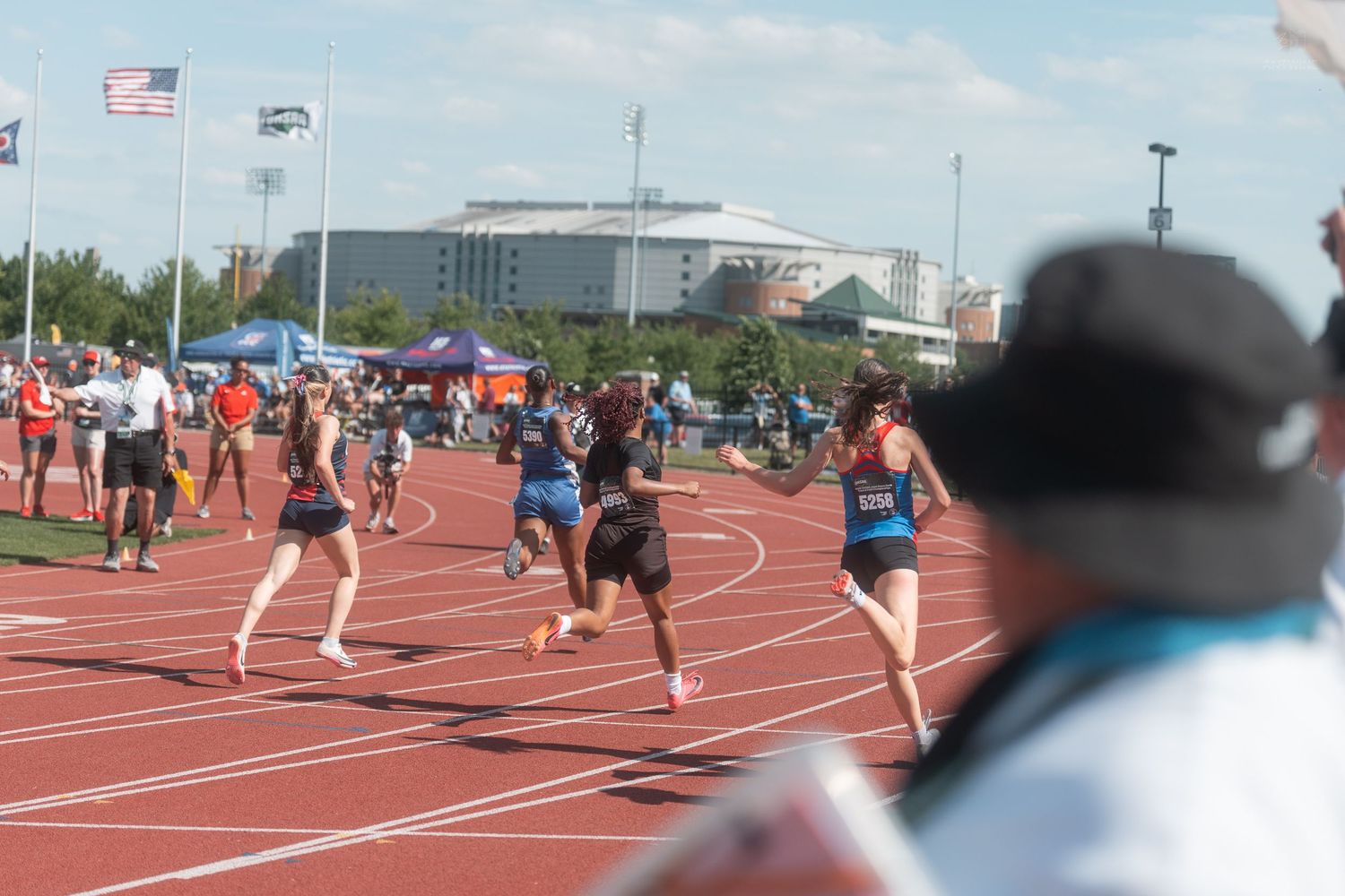 Runners compete in a relay race on a red track at an outdoor track and field competition with spectators watching.
