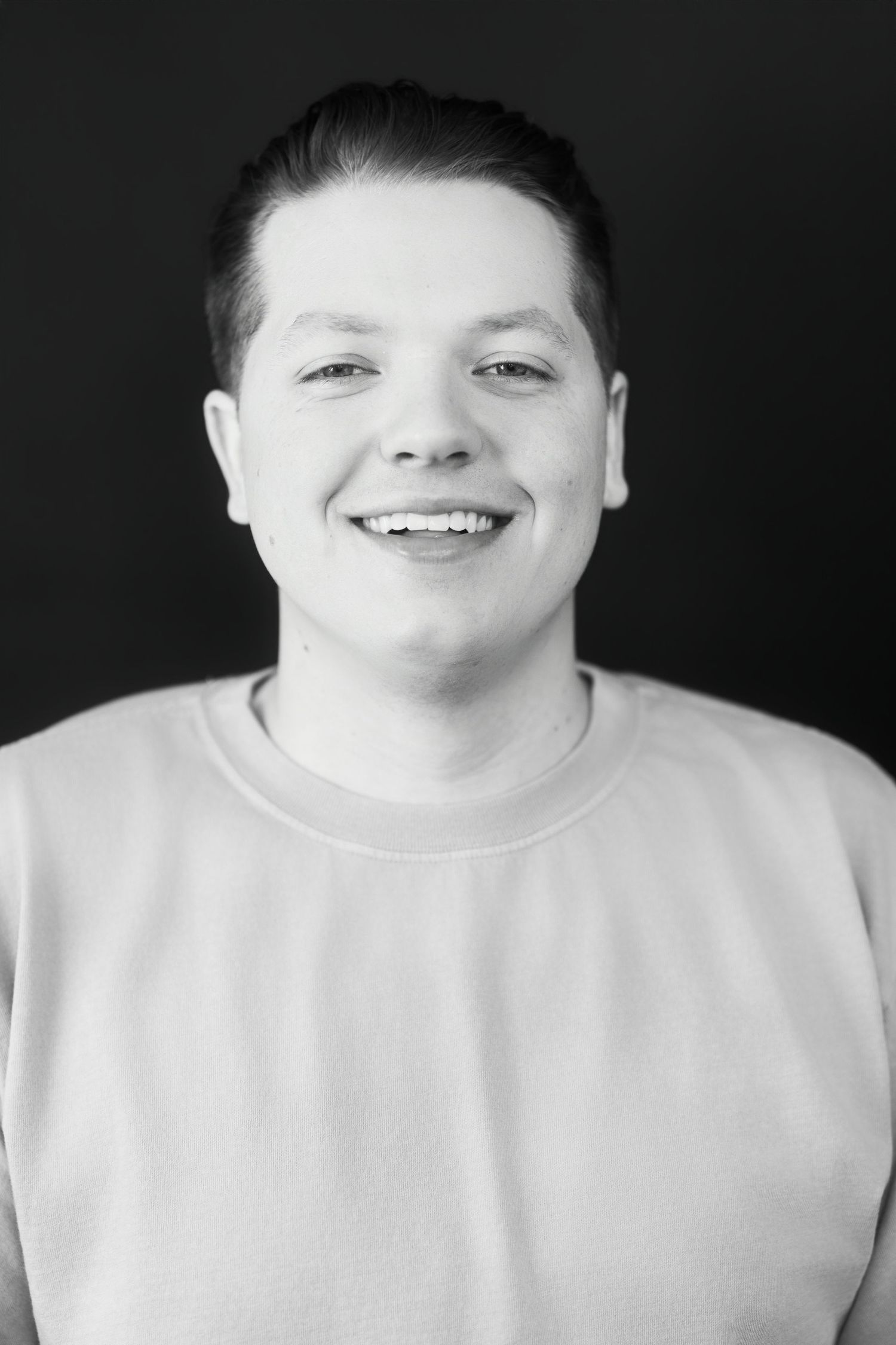 Black and white portrait of smiling young man in sweatshirt against dark background.