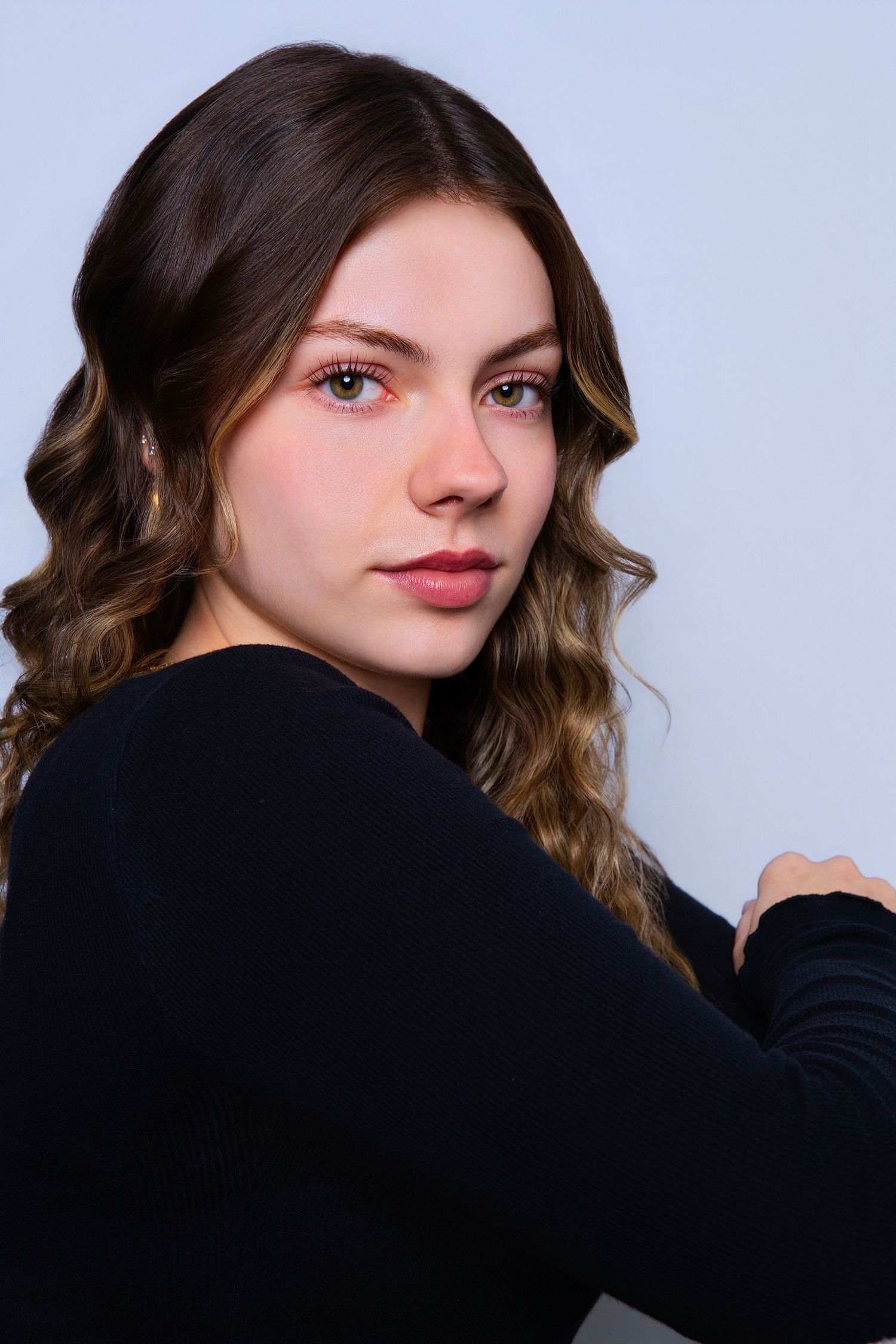 Young woman with curly highlighted hair in black long-sleeve top poses over shoulder.