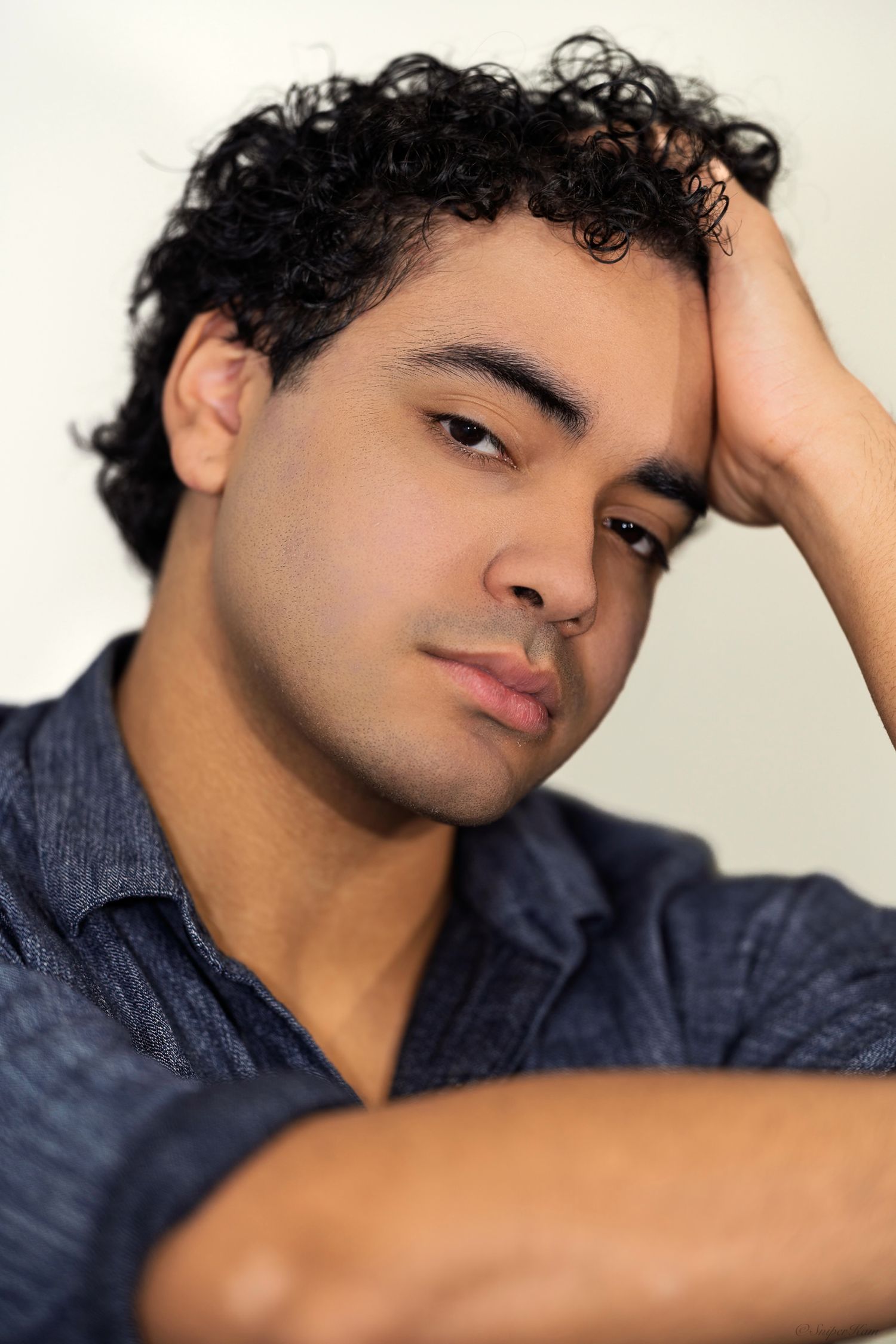Young man with curly black hair resting hand on head with thoughtful expression.