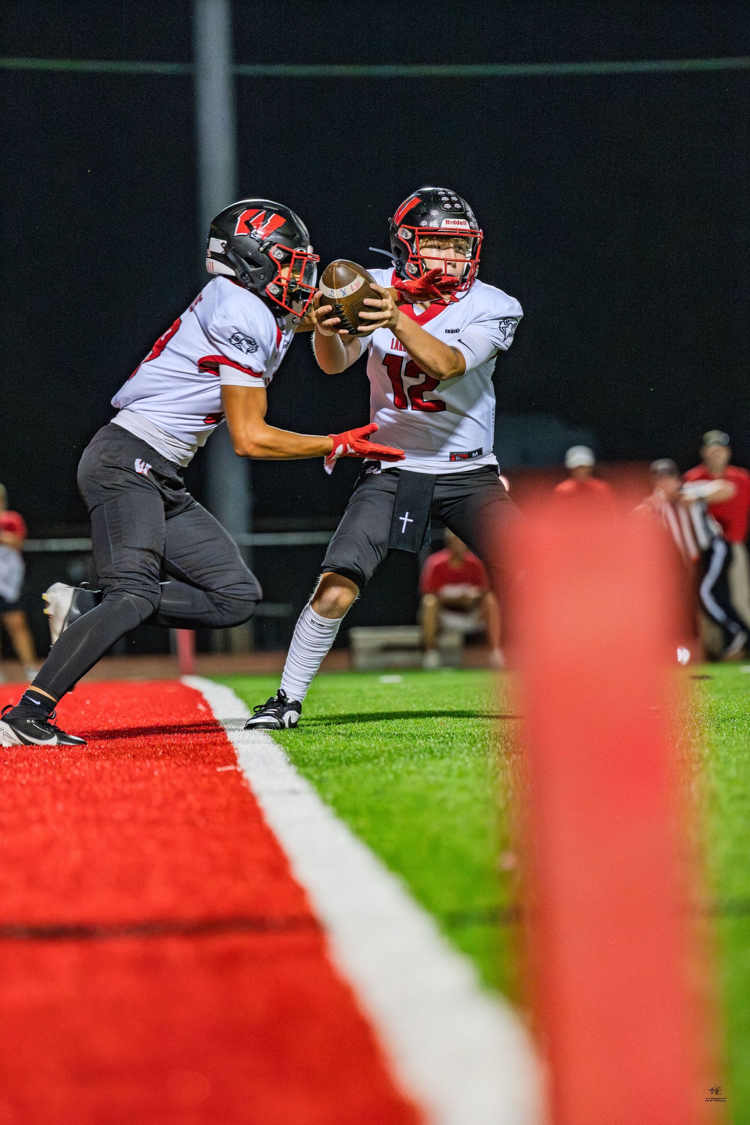 Football quarterback hands off ball to teammate near end zone during nighttime high school game.