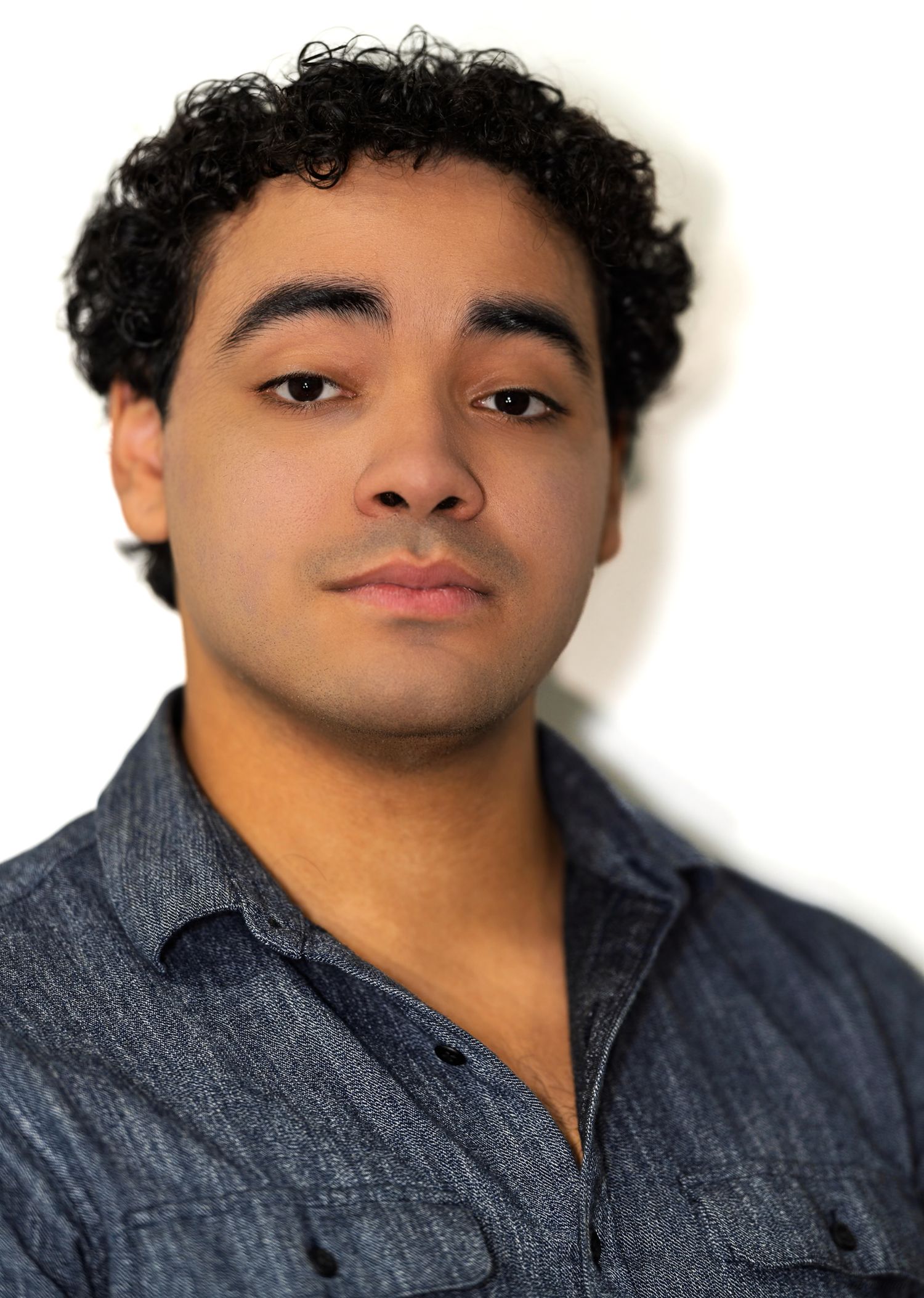 Young man with curly black hair wearing a blue denim shirt posing for a professional color headshot.