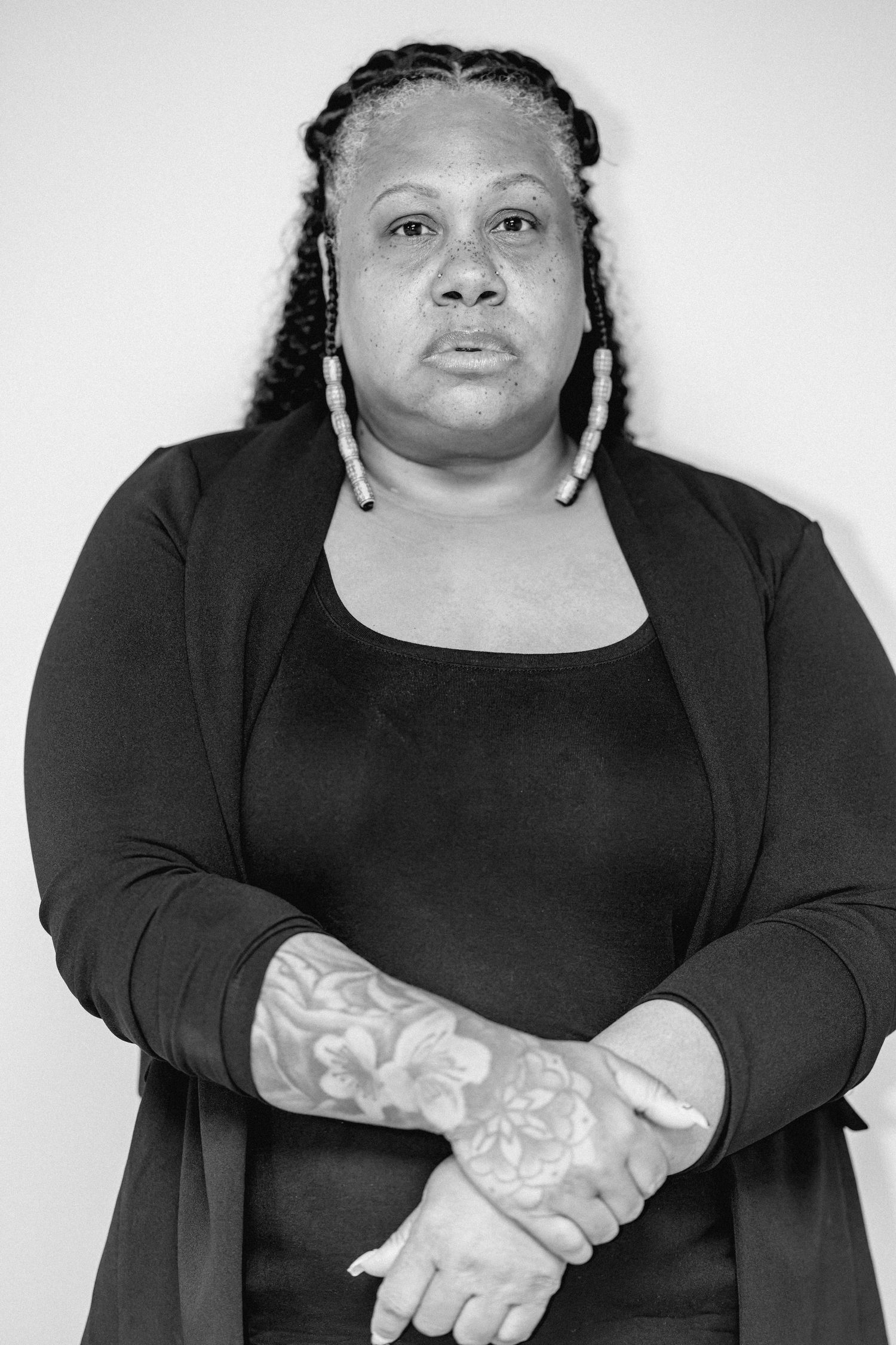 Black woman in black cardigan with floral arm tattoo, arms crossed, serious expression, monochrome photo.