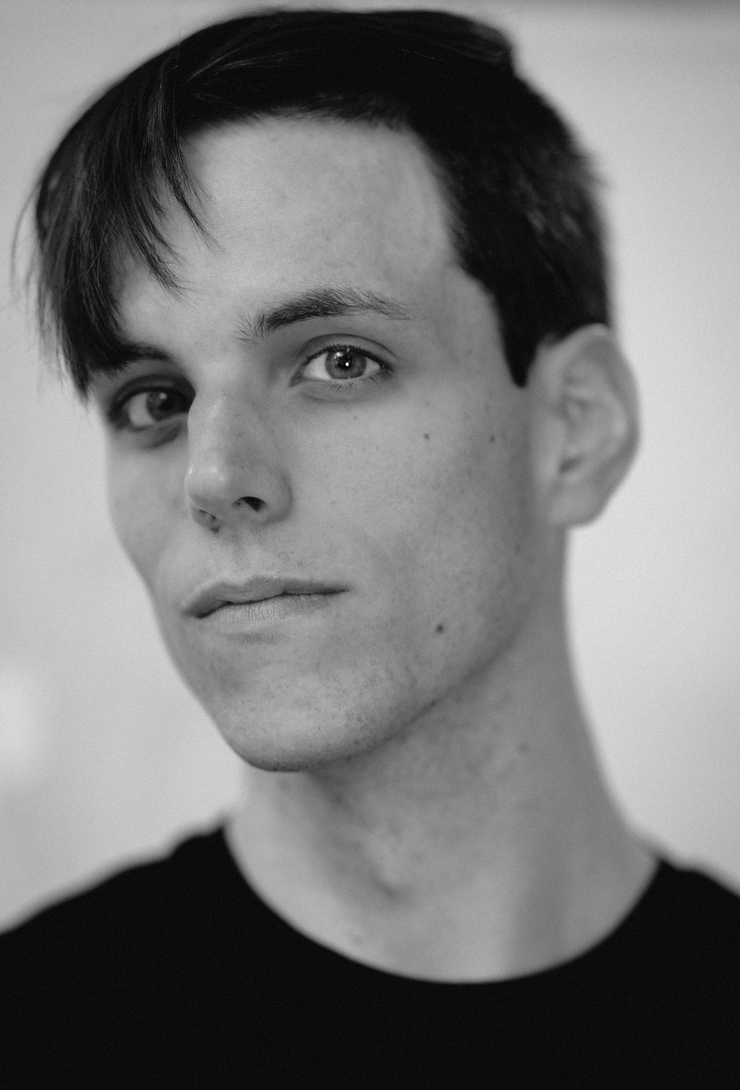 Black and white professional headshot of a young man with dark hair wearing a black t-shirt.