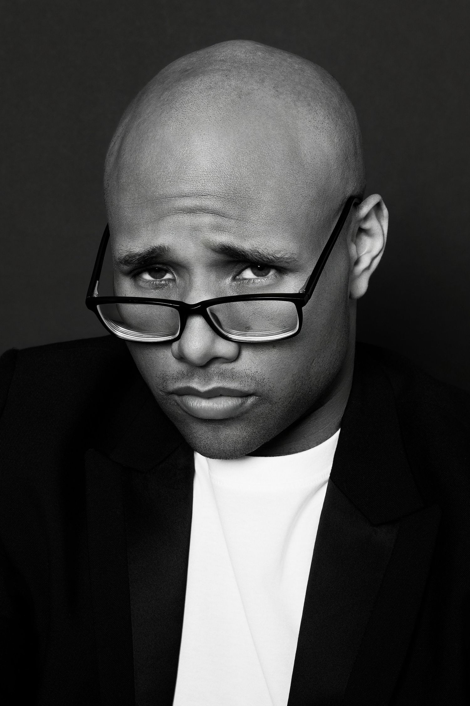Serious bald Black man in black blazer peering over rectangular glasses, dramatic black and white portrait photo.