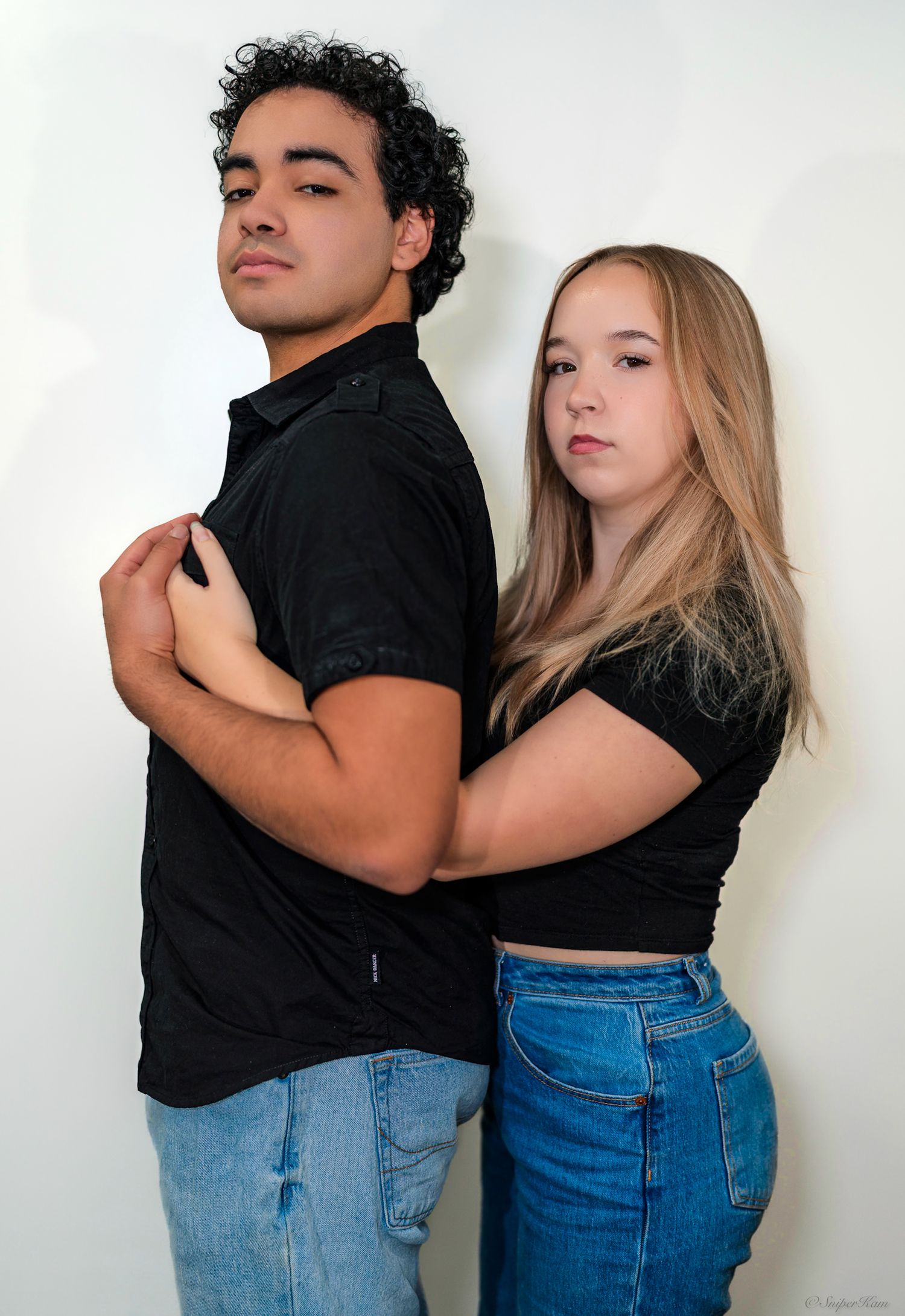 A young man and woman in matching black tops and jeans pose back-to-back against a white studio backdrop.