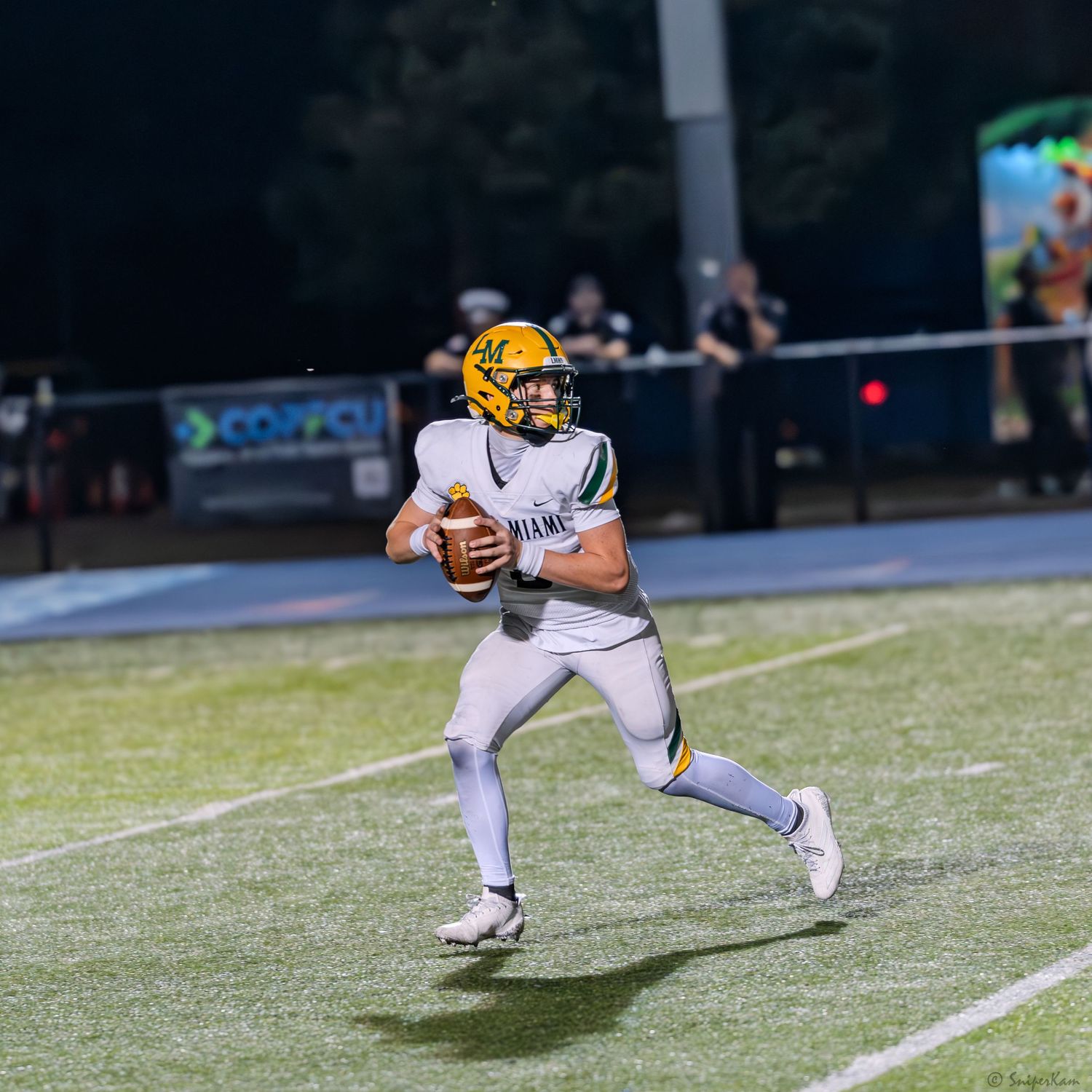 A quarterback in white and gold uniform drops back to pass during a nighttime high school football game.