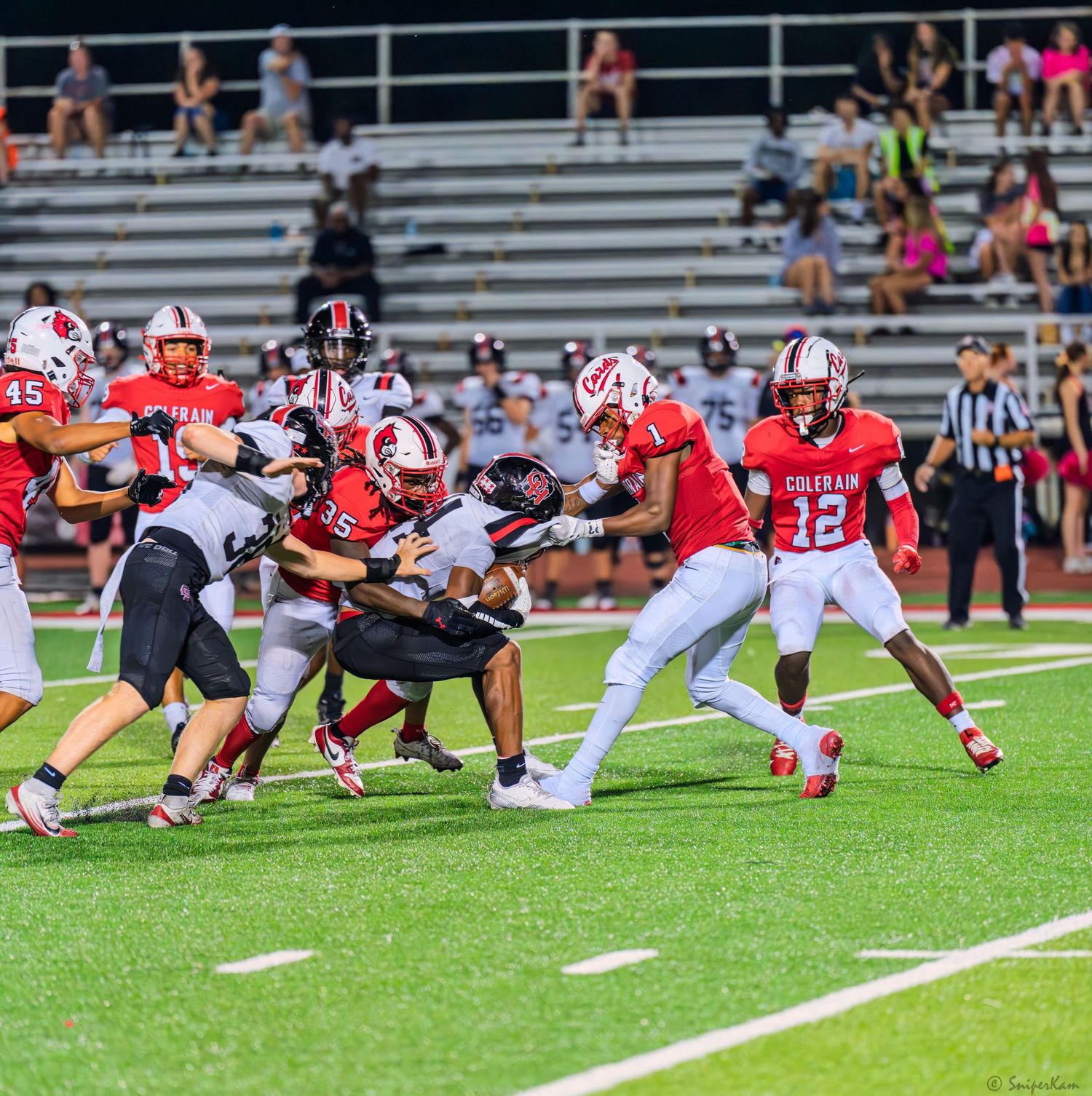 Colerain Cardinals players in red uniforms execute a running play against opponents during a home game.