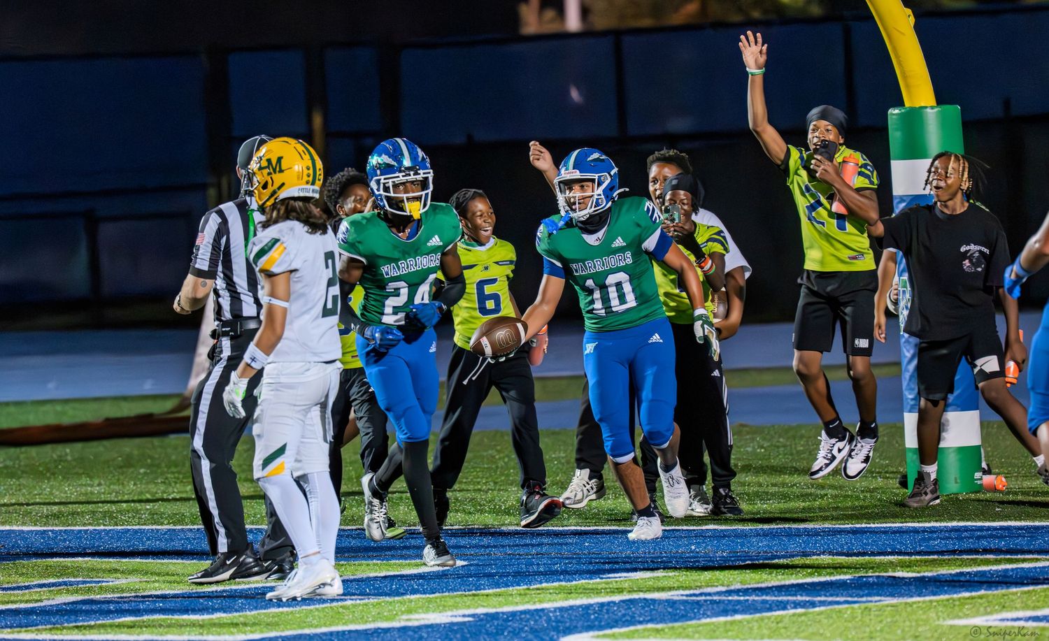 High school football players in green and blue uniforms celebrate touchdown with referee signaling score.