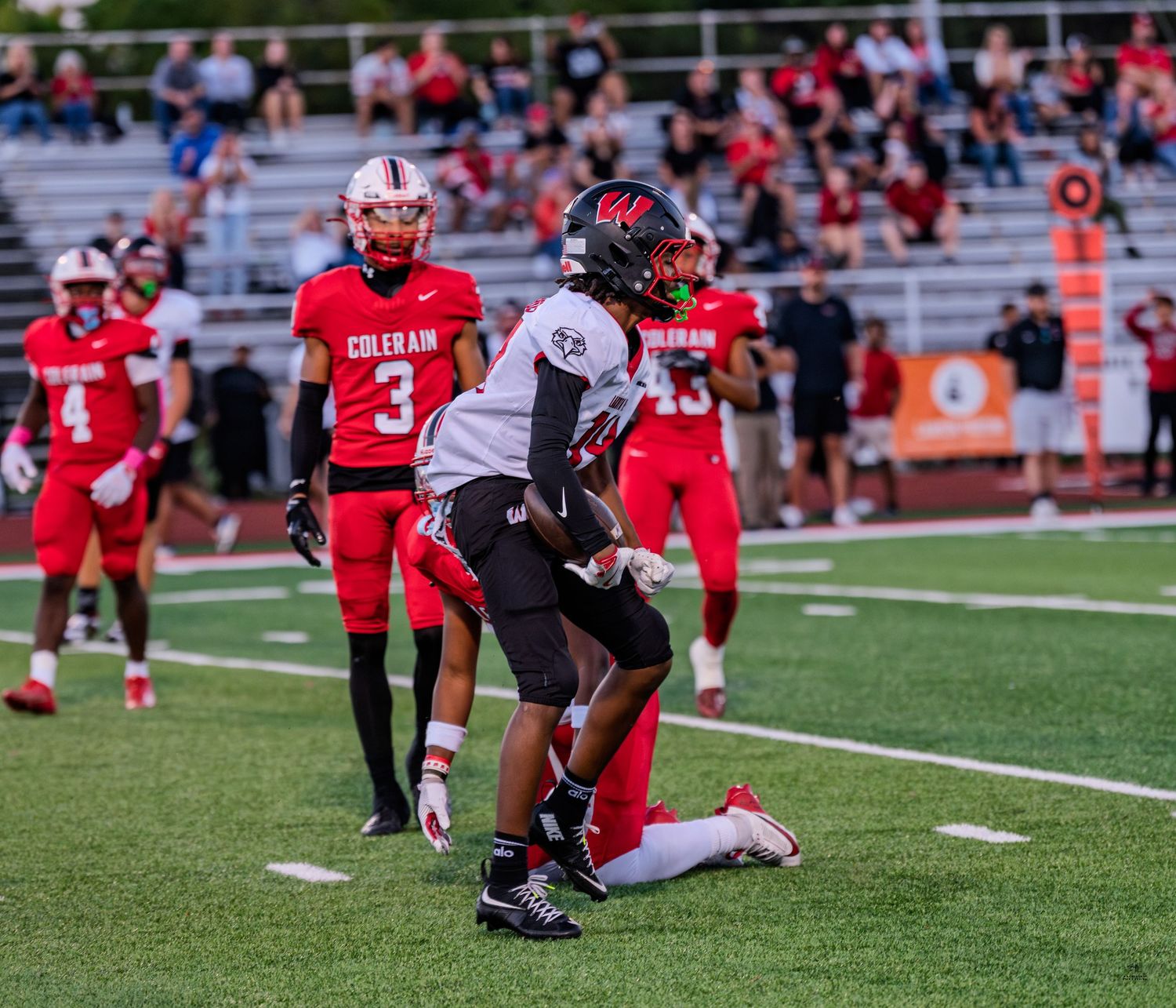 Football player kneels on field during high school game while Colerain Cardinals players stand in background.