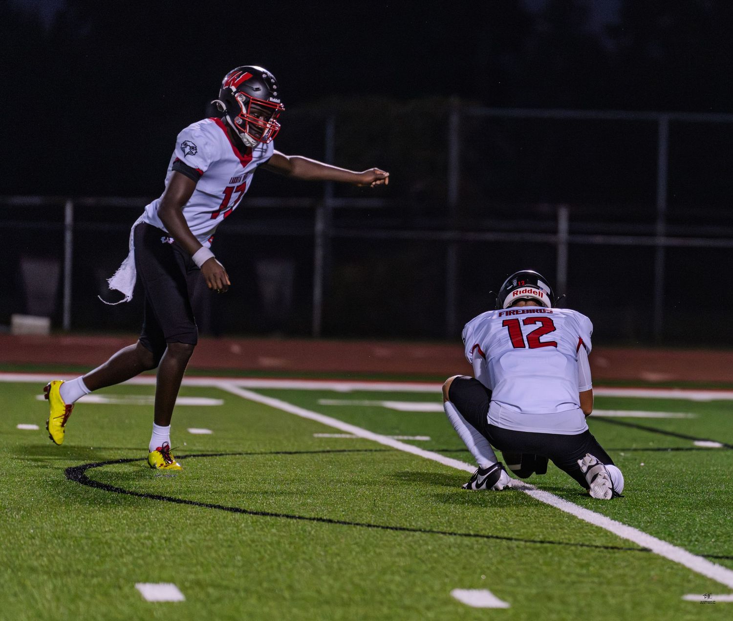 A football player kicks a field goal attempt with a holder kneeling during a night high school game.