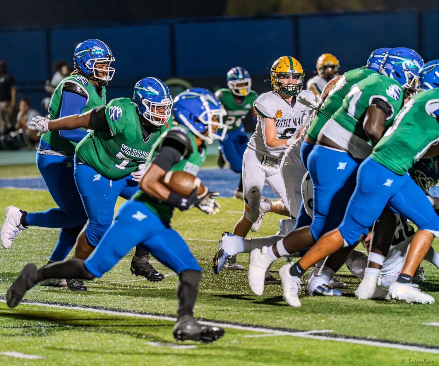High school football players in green and blue uniforms execute a play during a night game under stadium lights.