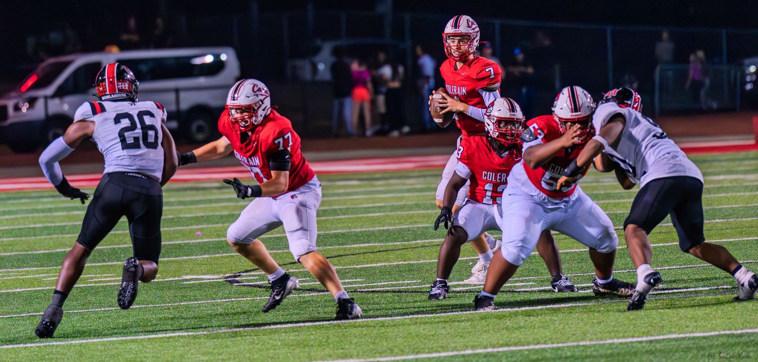 Red-uniformed football players execute a running play as a defender in black jersey #26 pursues during a high school game.