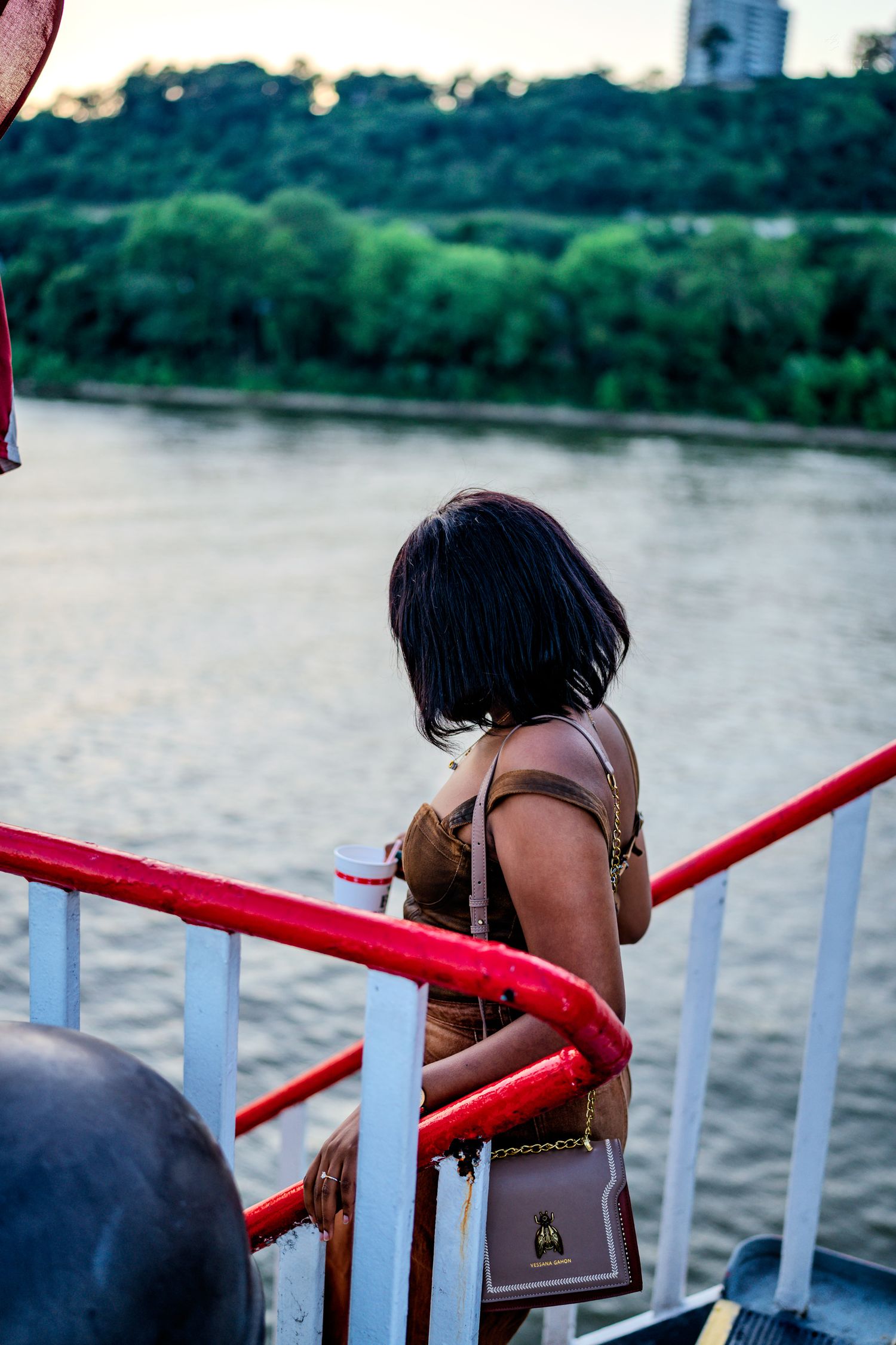 Woman with black bob hair and brown bag standing at red railing overlooking a river at dusk.