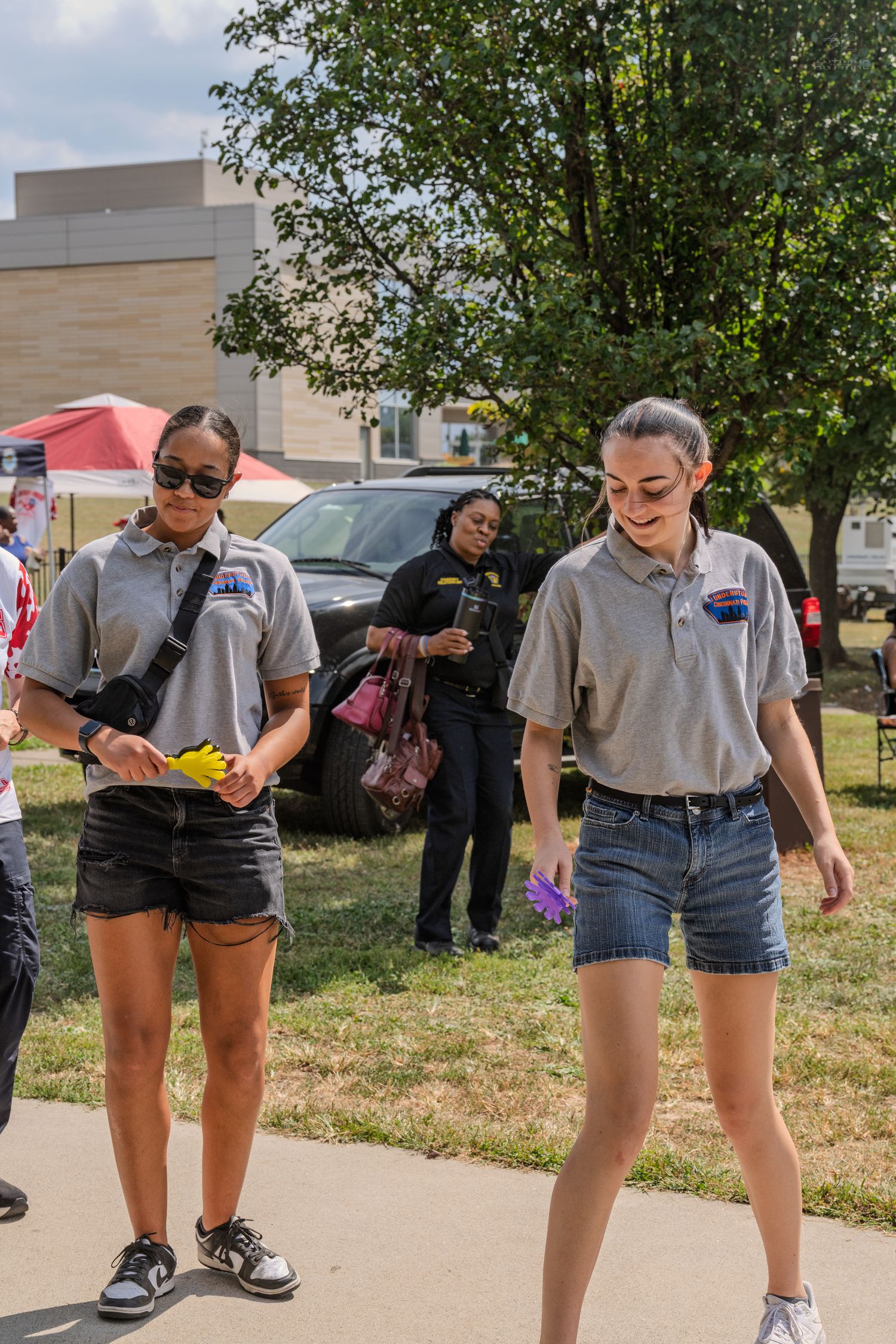 Two young women in matching gray polo shirts walk together at an outdoor community sports event.