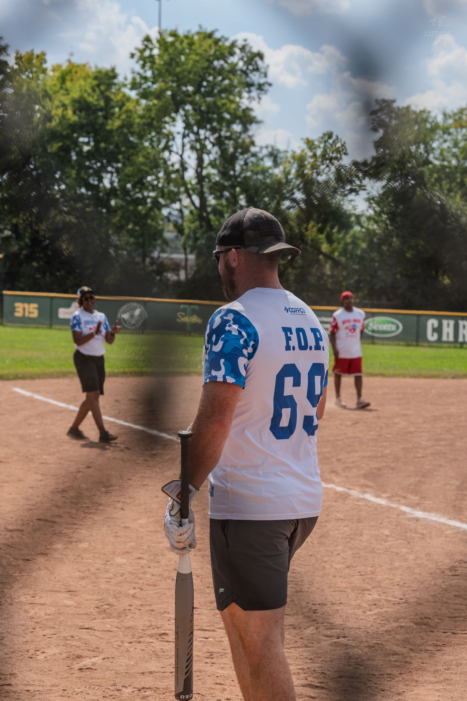 Softball player wearing F.O.R. jersey number 69 holding a bat while watching teammates play on a sunny field.
