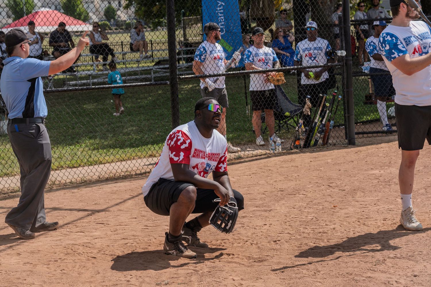Softball catcher crouching in position on dirt field during sunny outdoor charity game.