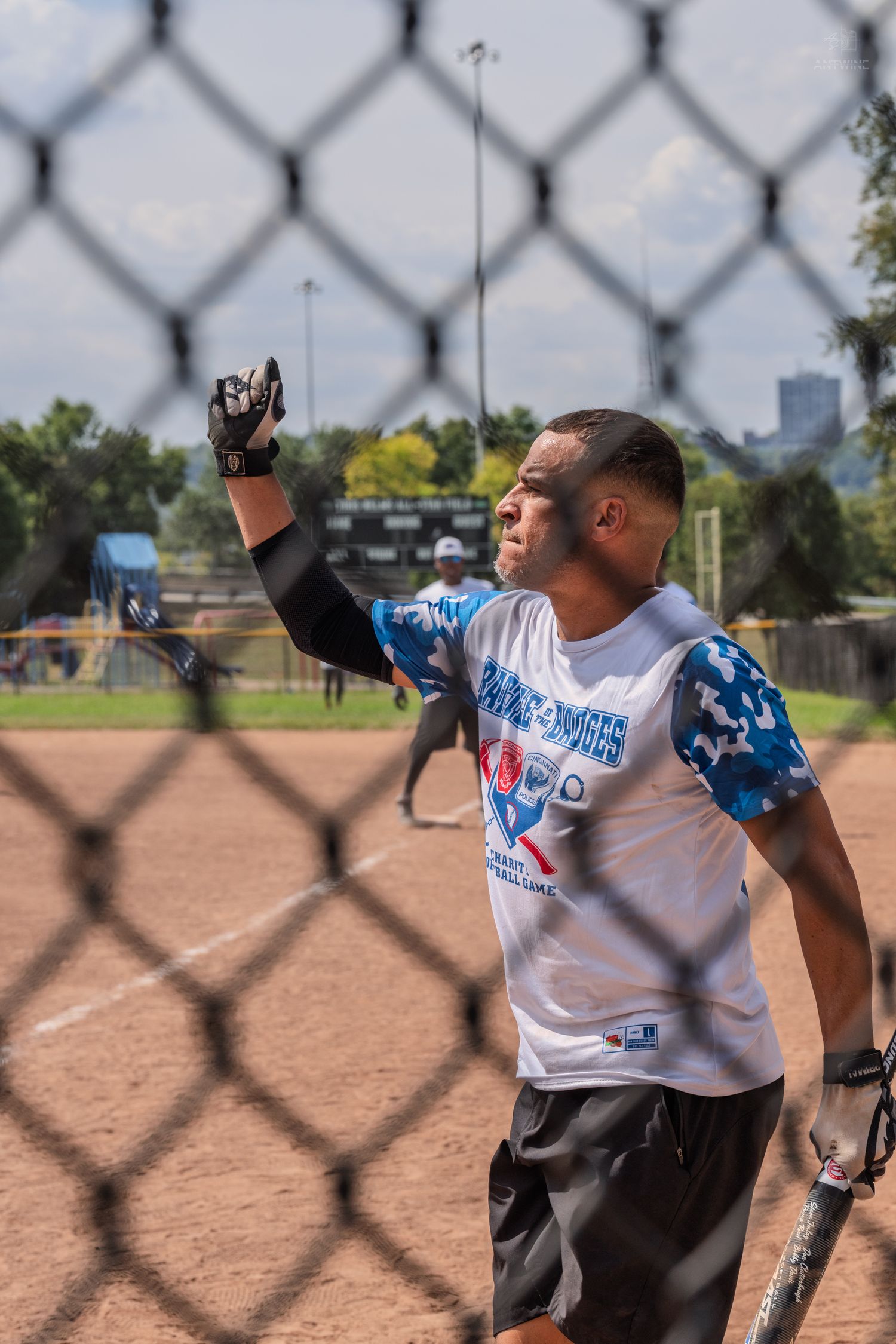 Softball player viewed through chain-link fence raising glove during Battle of the Badges charity game.
