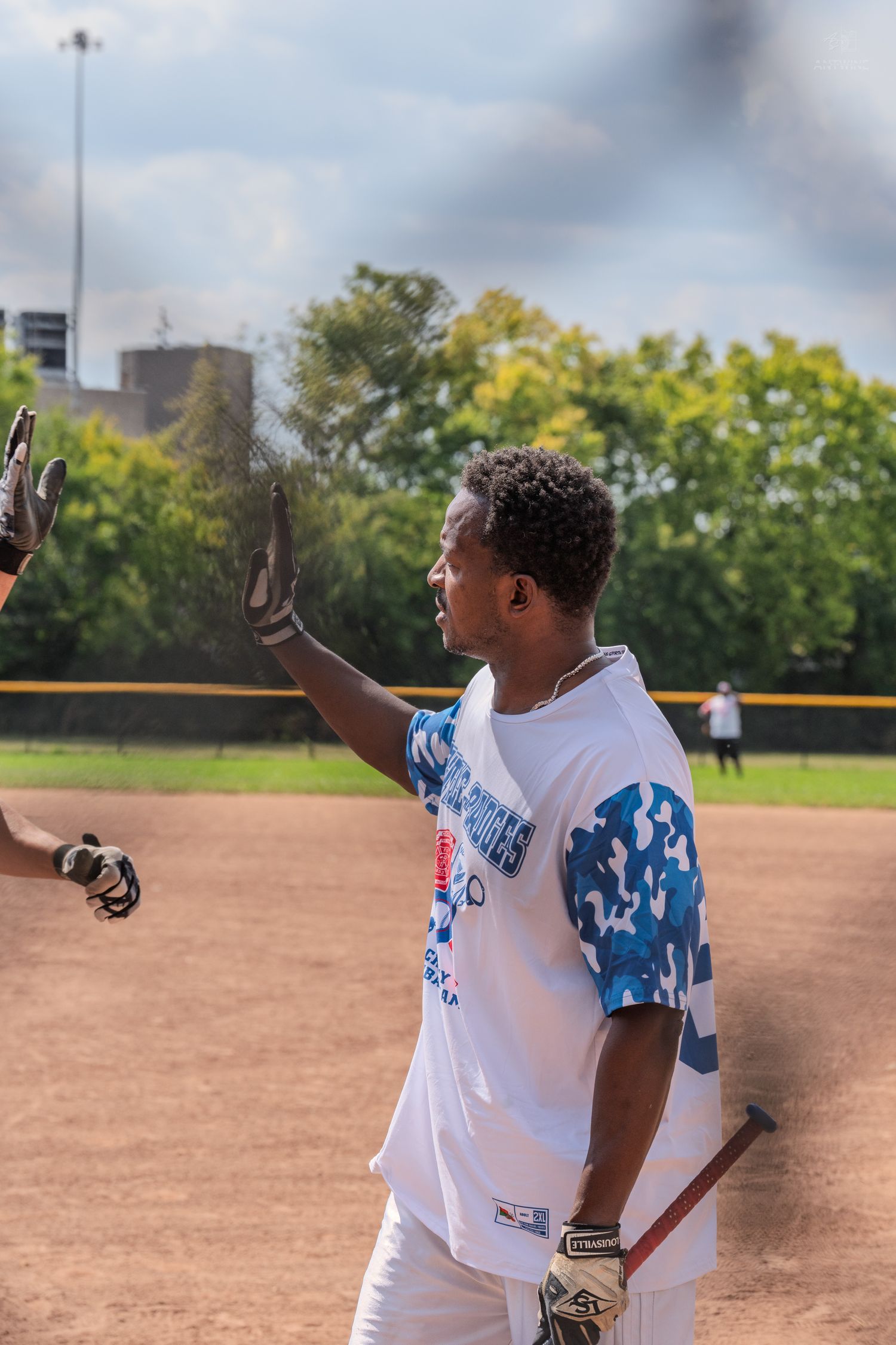 Young baseball player gives a high five wearing batting gloves and a blue camo jersey on a softball field.
