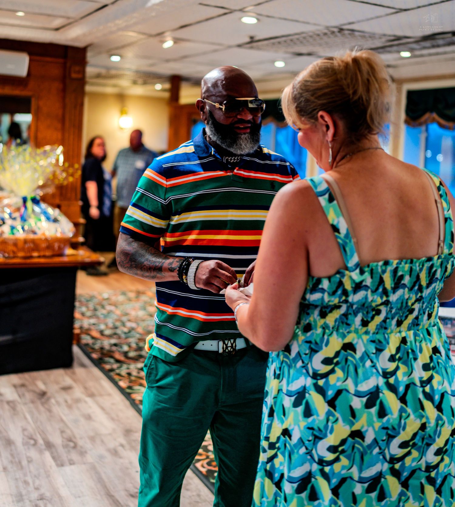 Two people conversing indoors at a casual event, man in striped polo and green pants, woman in teal floral dress.