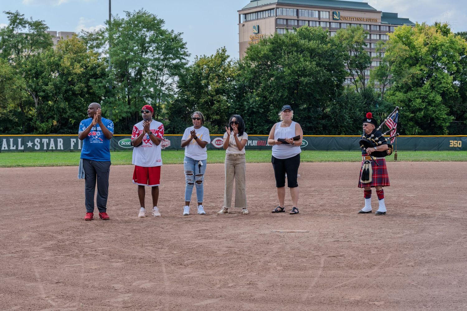 Seven people including a bagpiper lined up on an All-Star softball field during an opening ceremony.