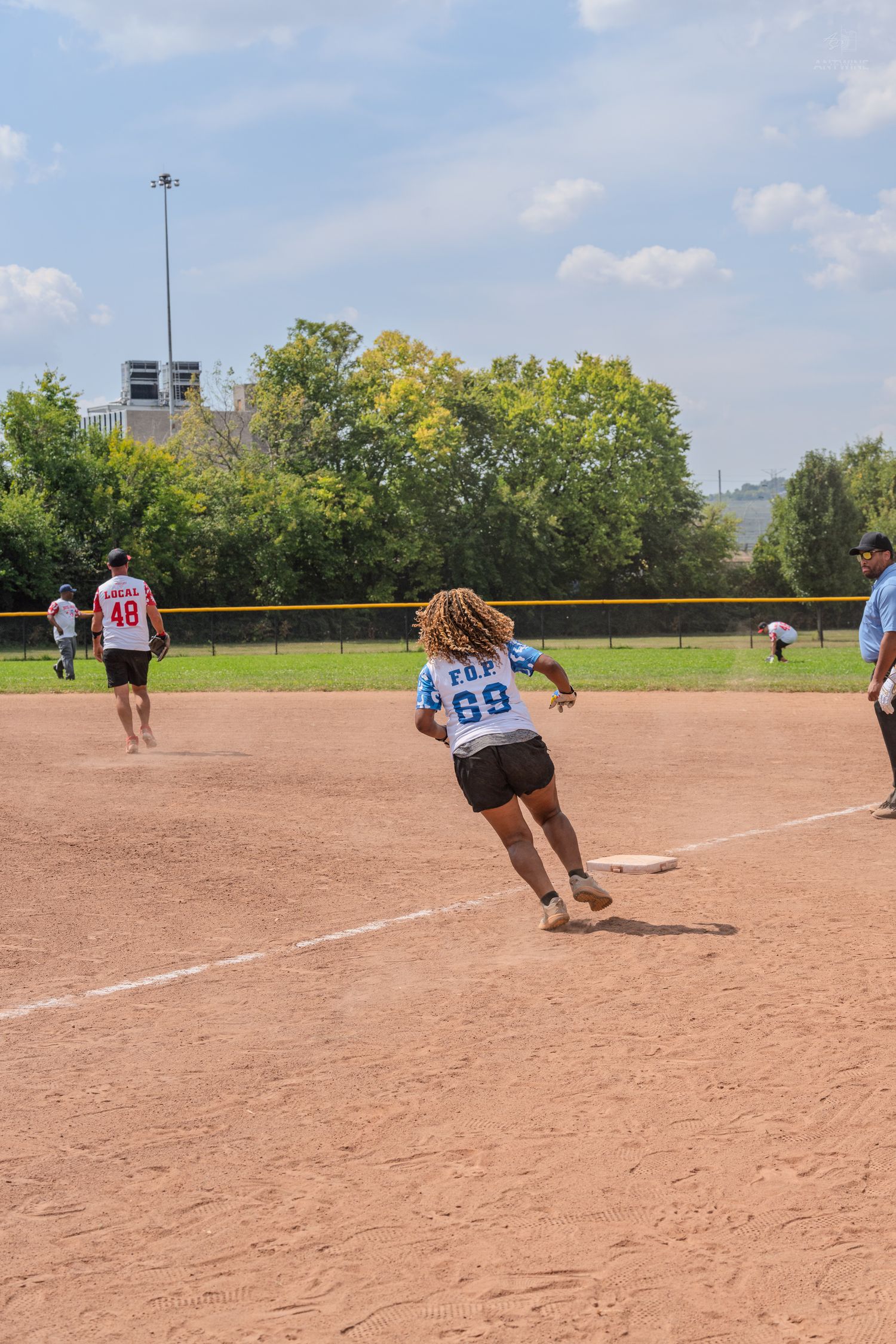 Player in jersey number 69 rounds a base during an outdoor softball game on a sunny day.