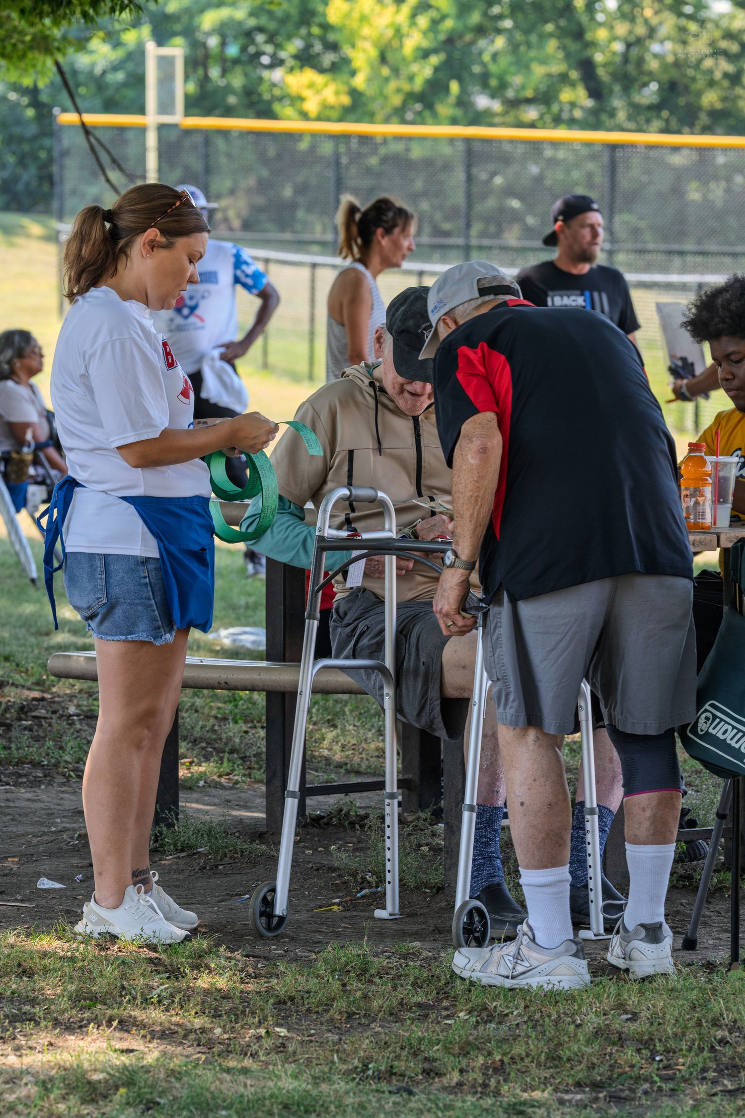 Volunteers assist an elderly man with a walker at an outdoor community event near a baseball field.