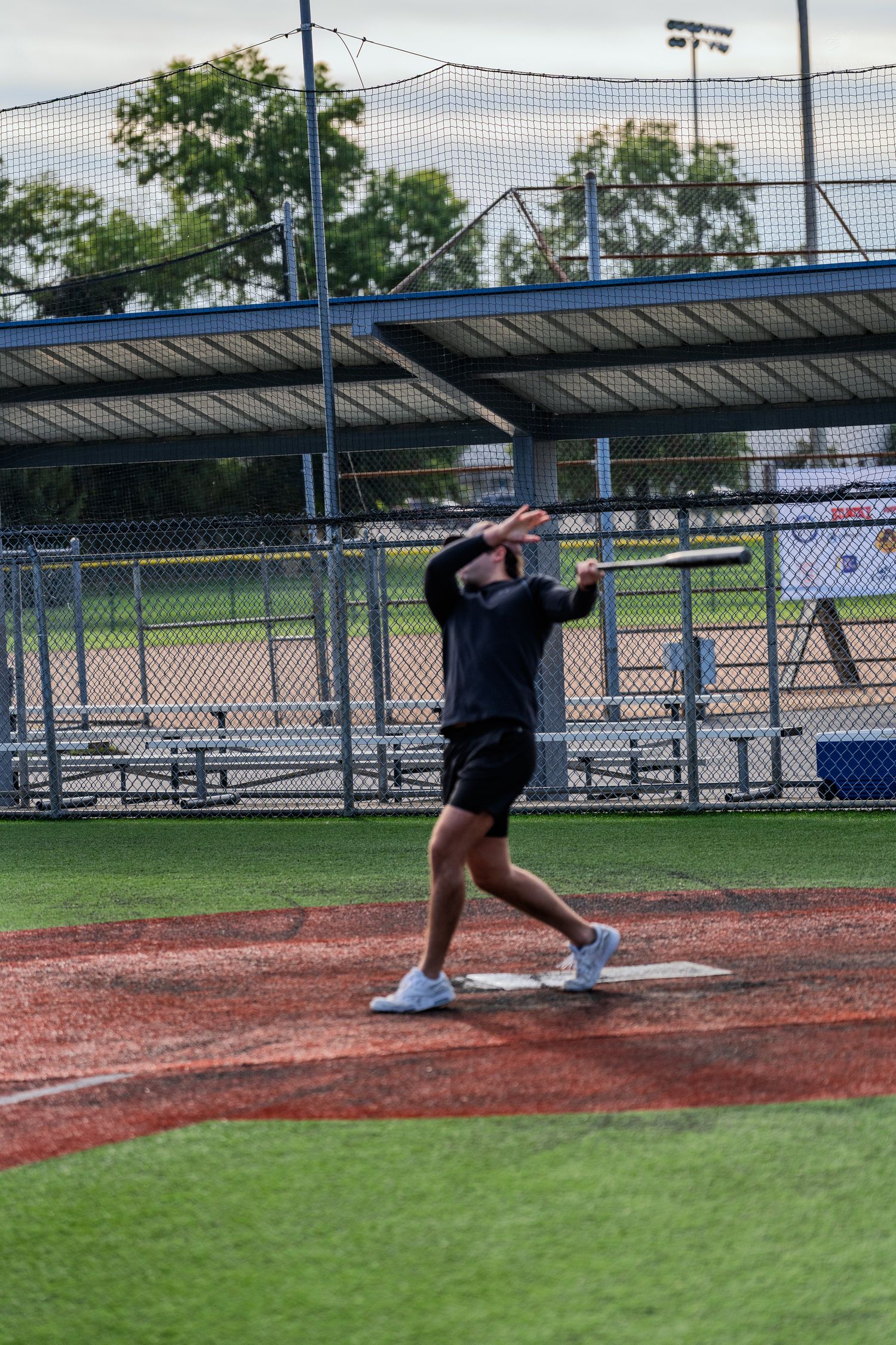 A baseball player swings a bat on a red dirt field at an outdoor baseball diamond with green turf.