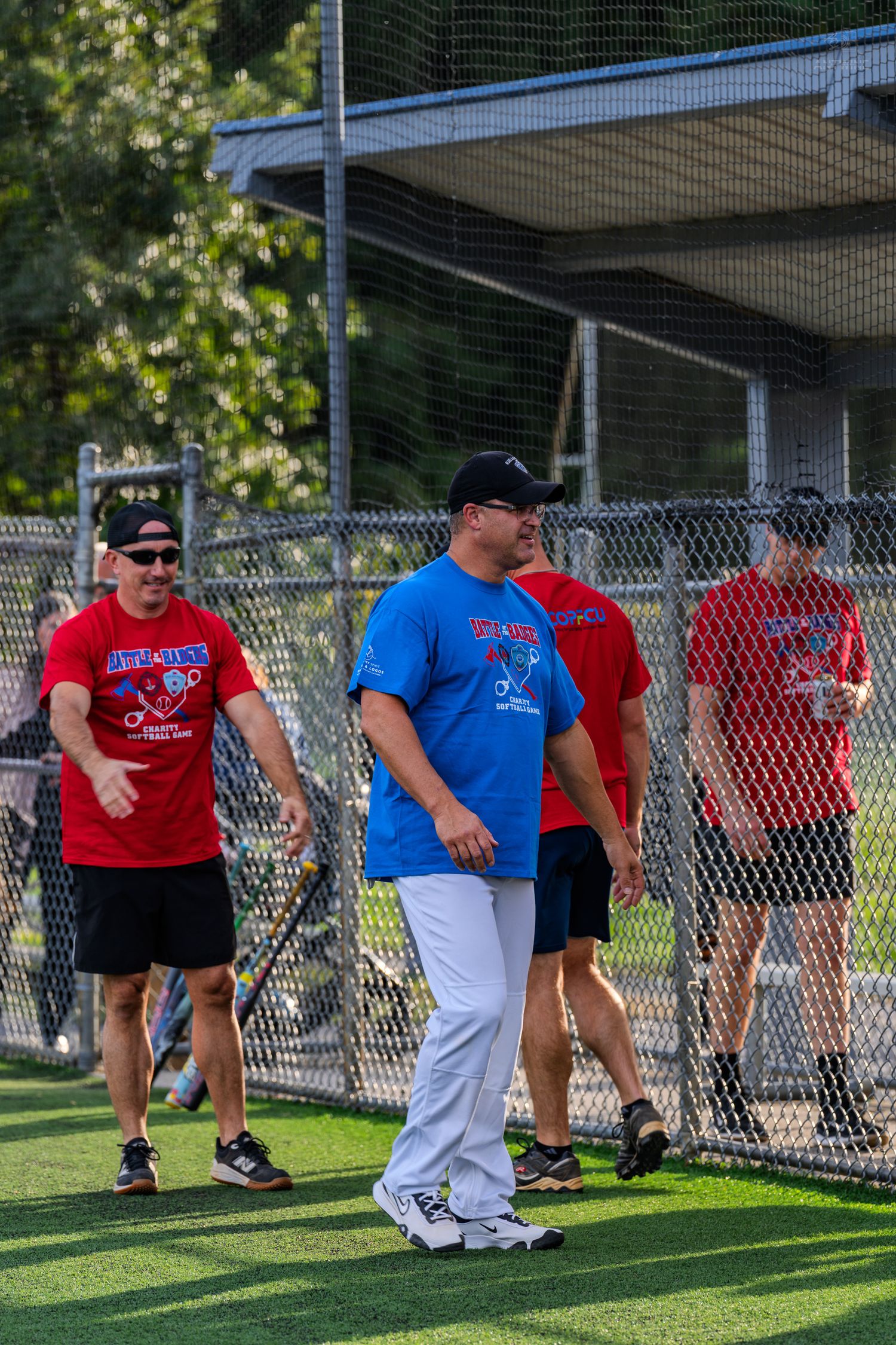 Men in red and blue charity softball game shirts walk near a chain-link fence on a green turf field.