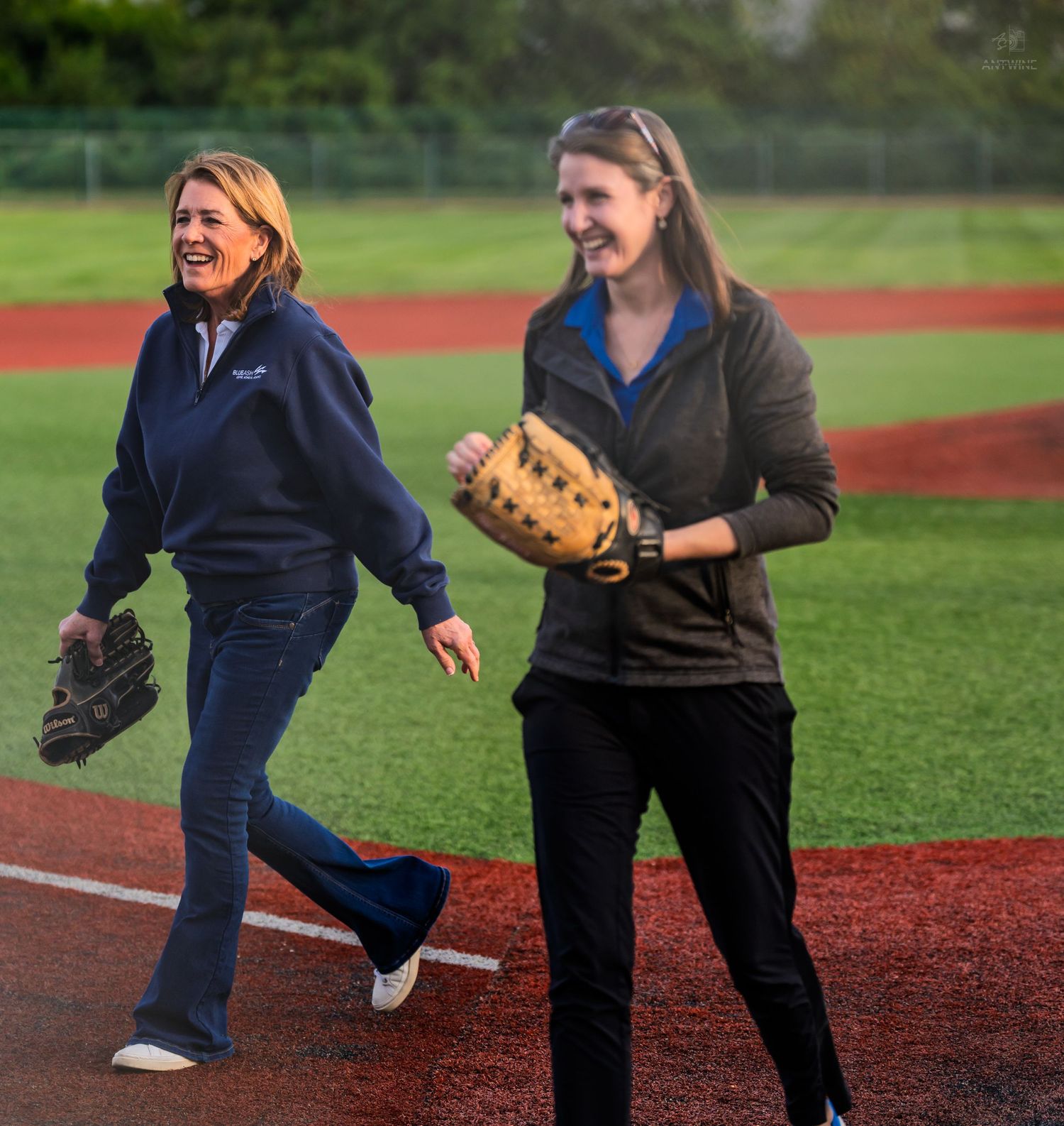Two women wearing baseball gloves laugh and walk together on a softball field during an outdoor game.