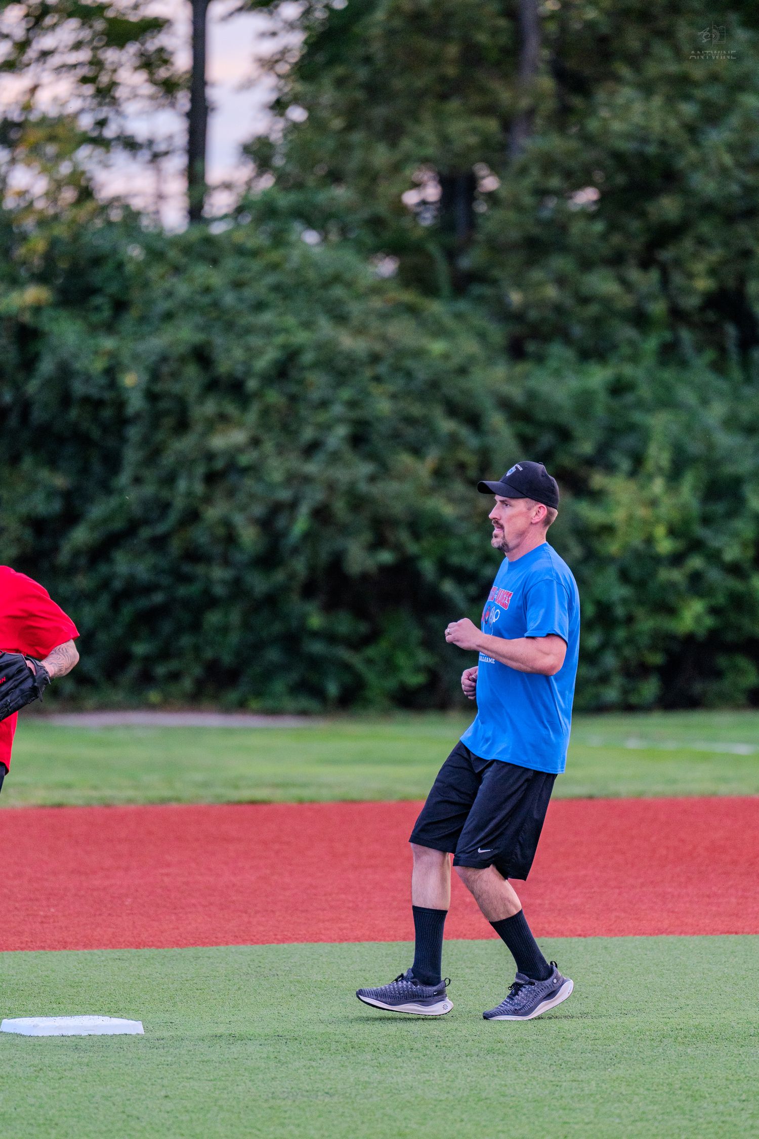 Male softball player in blue shirt and black shorts walking on field during an evening recreational game.
