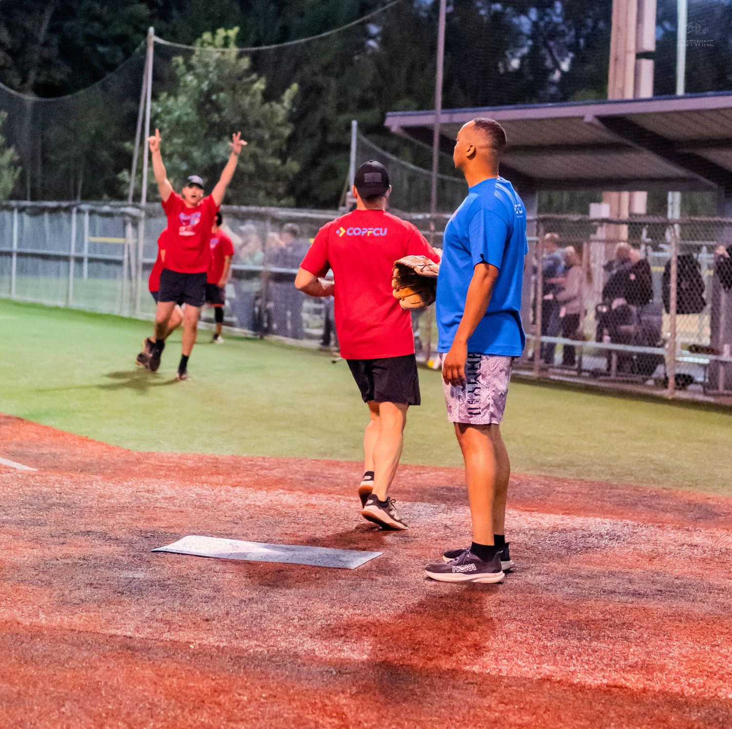 Softball players celebrate near home plate as a teammate scores a run during a recreational league night game.