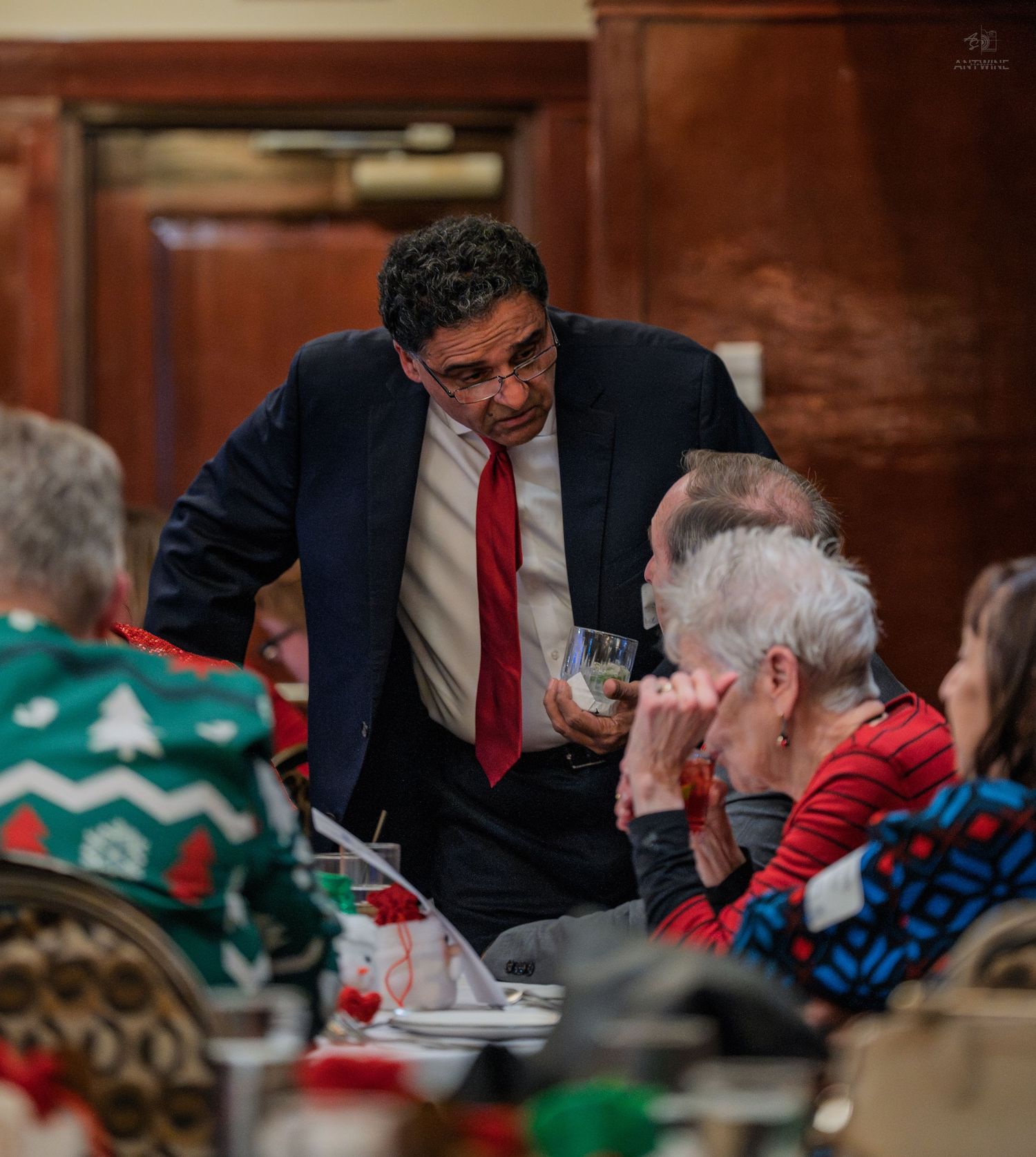 Man in suit with red tie speaks to elderly guests at a festive Christmas dinner party in elegant dining room.