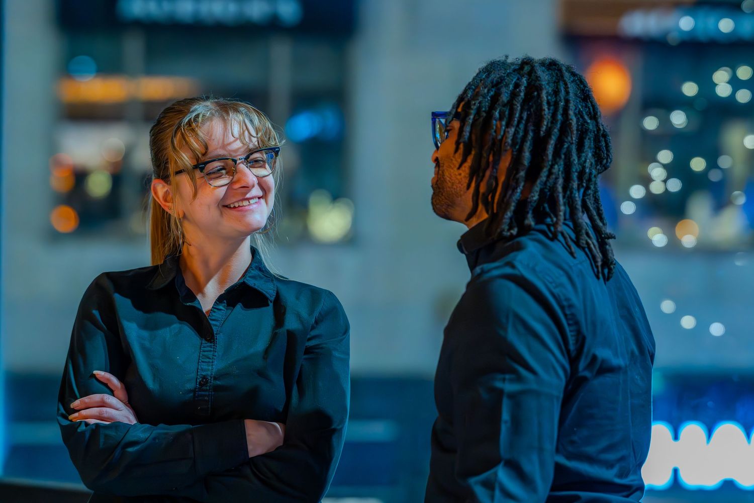 Two professionals in black shirts engage in conversation outdoors at night with bokeh city lights in background.