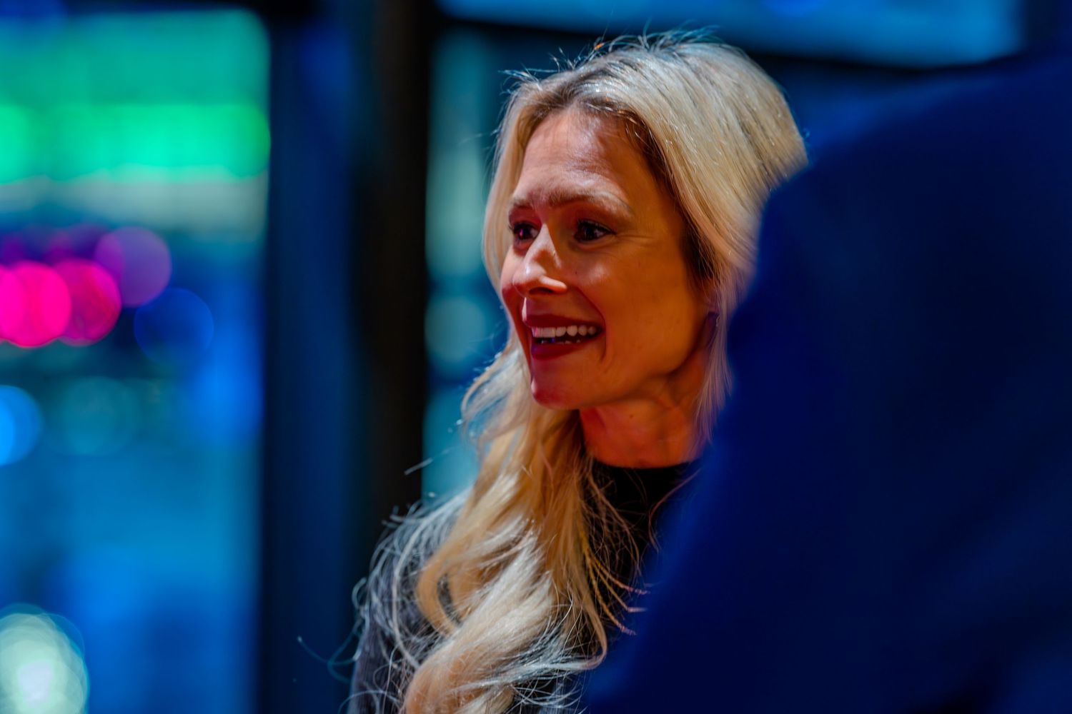 Blonde woman smiling in dark setting with colorful lights in background, wearing navy blue top.