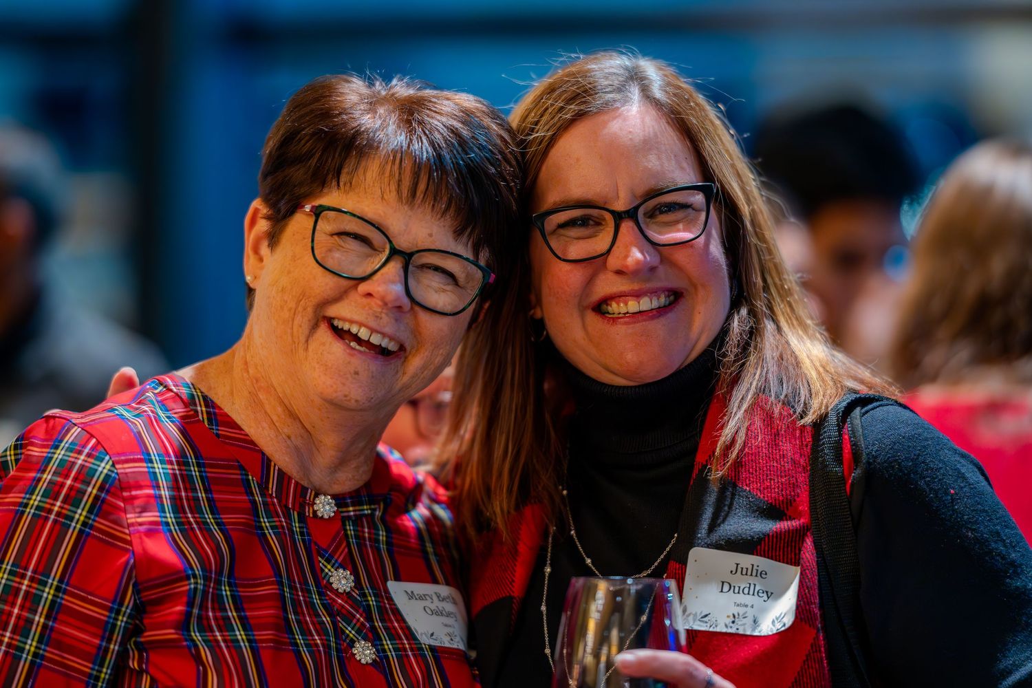 Two smiling women wearing glasses and red outfits pose cheek-to-cheek at a festive holiday event.