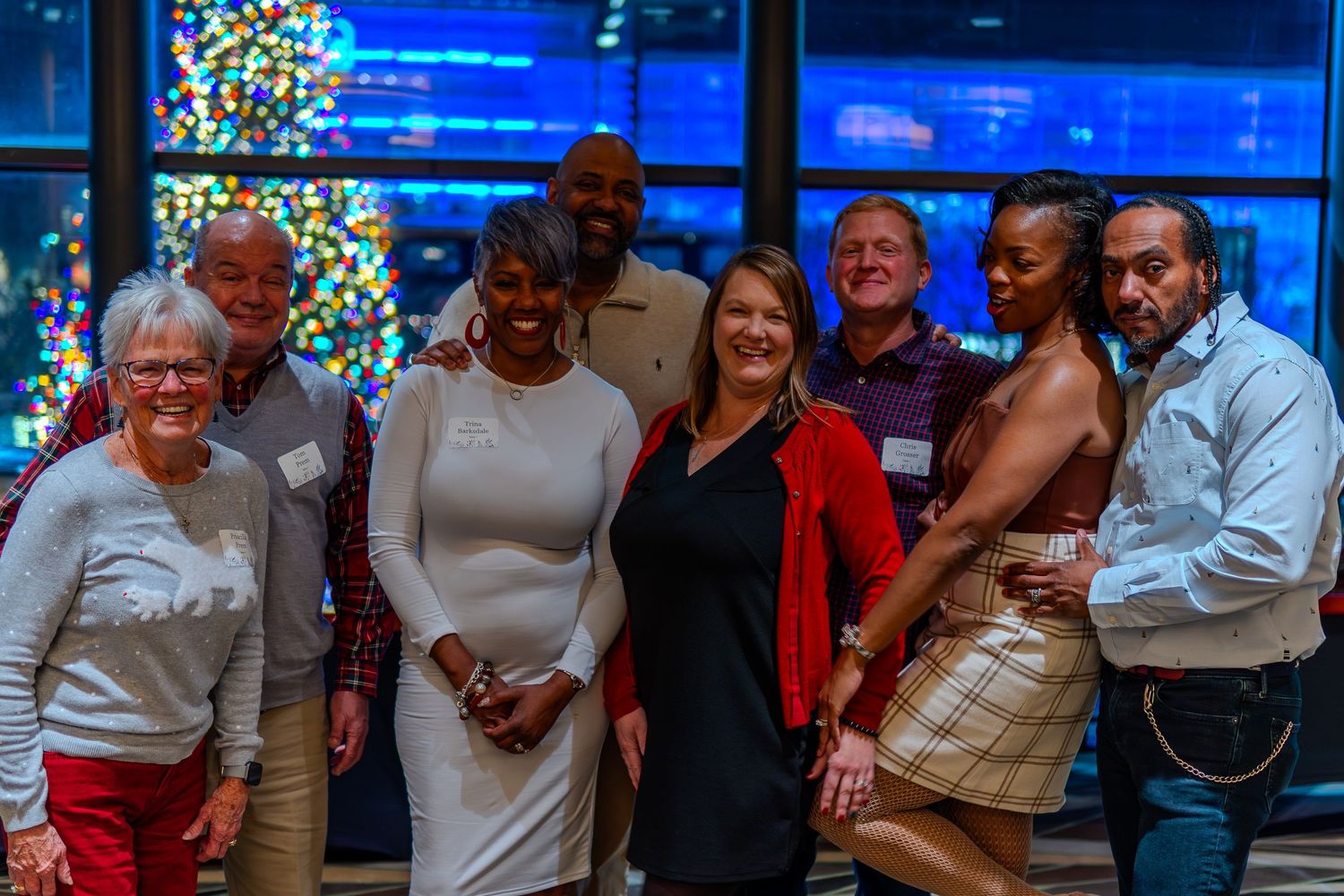 Group of nine people smiling together at a festive holiday party with a decorated Christmas tree in the background.