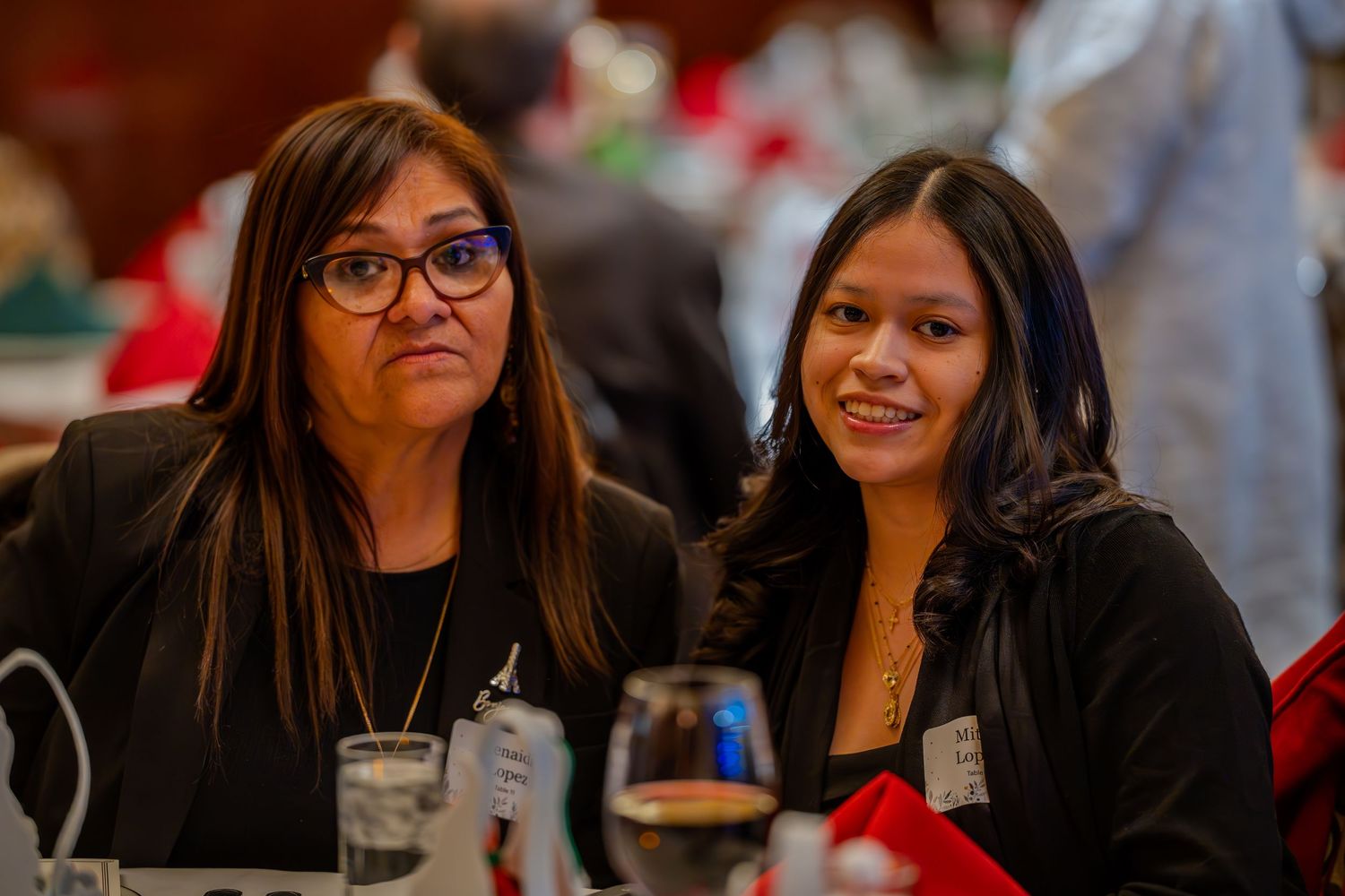 Two women in black blazers smile at a formal dinner event, seated at a table with glasses and decorations.