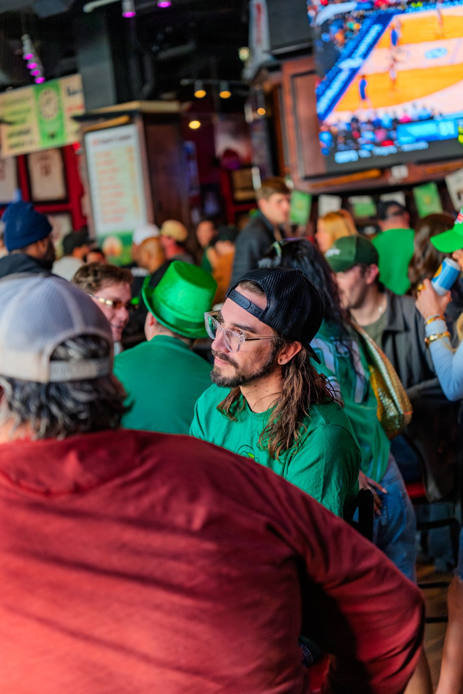 Crowd celebrating St. Patrick's Day at a bar, wearing green hats and shirts, with a basketball game on TV.