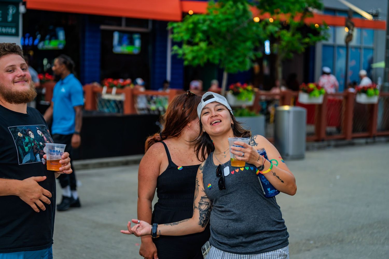 Two women laughing and dancing outdoors at a lively street festival, holding drinks near a colorful bar patio.
